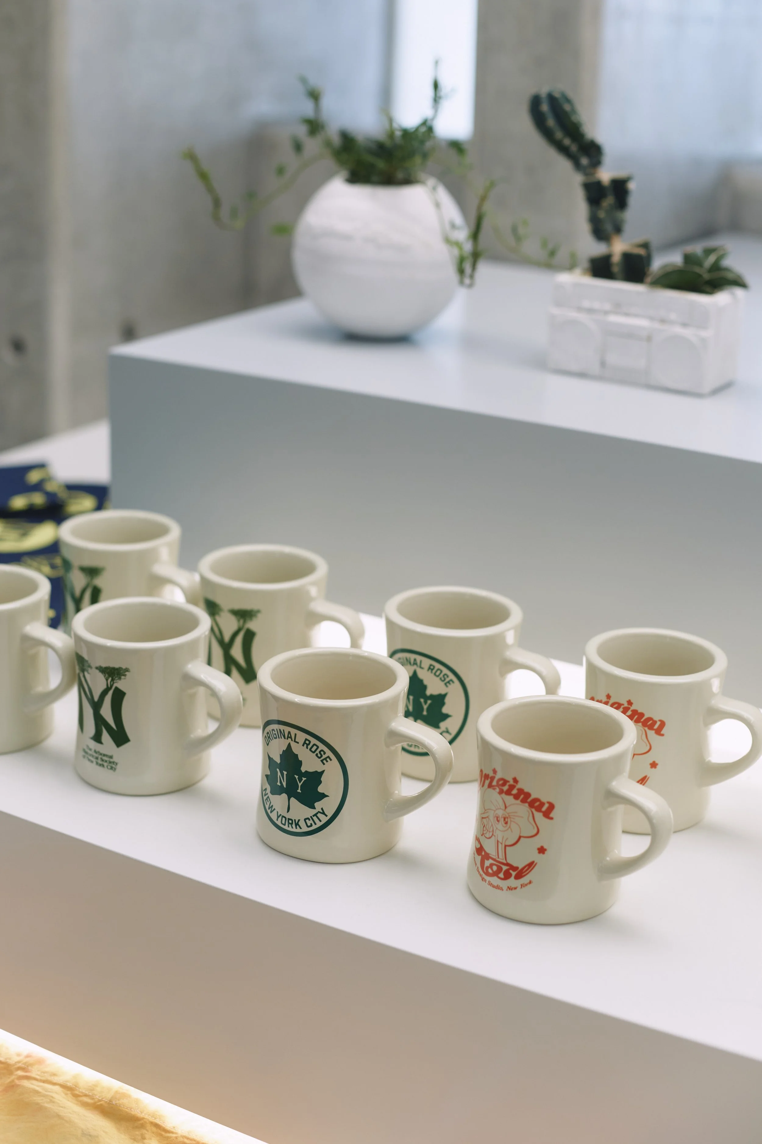 Multiple New York City souvenir mugs on a white display table, with a white vase with green plants and a white rectangular container with a plant in the background.