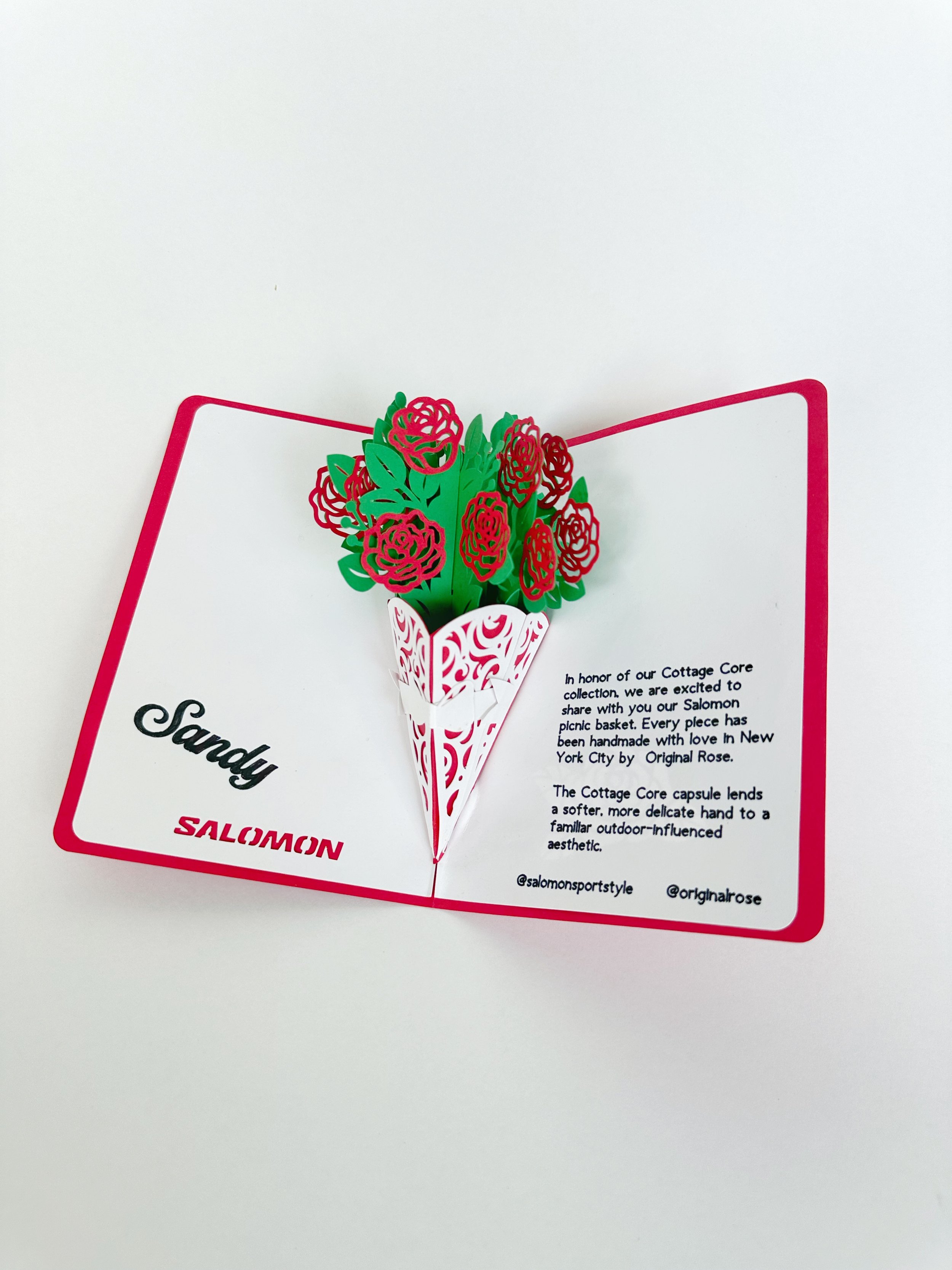 A pop-up card with a 3D paper bouquet of red and pink roses and green leaves, displayed open on a white background. The card features the words "Sandy" in black script and "SALMON" in red block letters, with printed text about the Cottage Core collec
