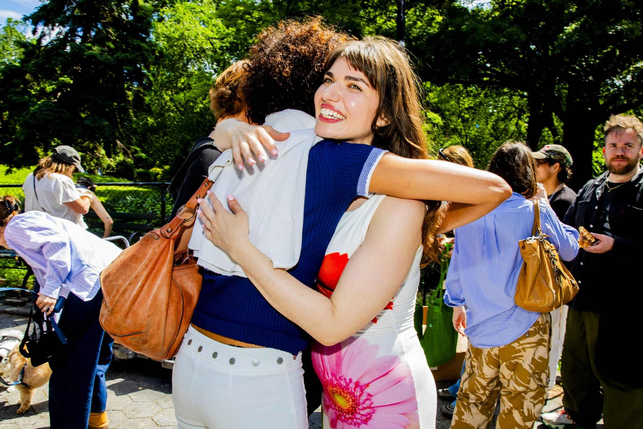 Two women hugging each other and smiling, surrounded by other people outdoors on a sunny day in a park.