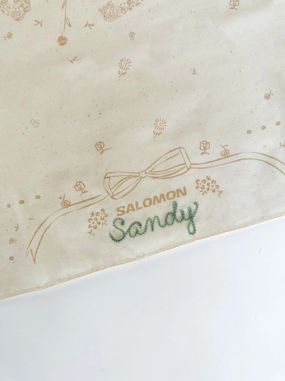Close-up of a decorative piece with the words 'SALOMON Sandy' and a bow design, featuring small flower and star patterns on a light background.