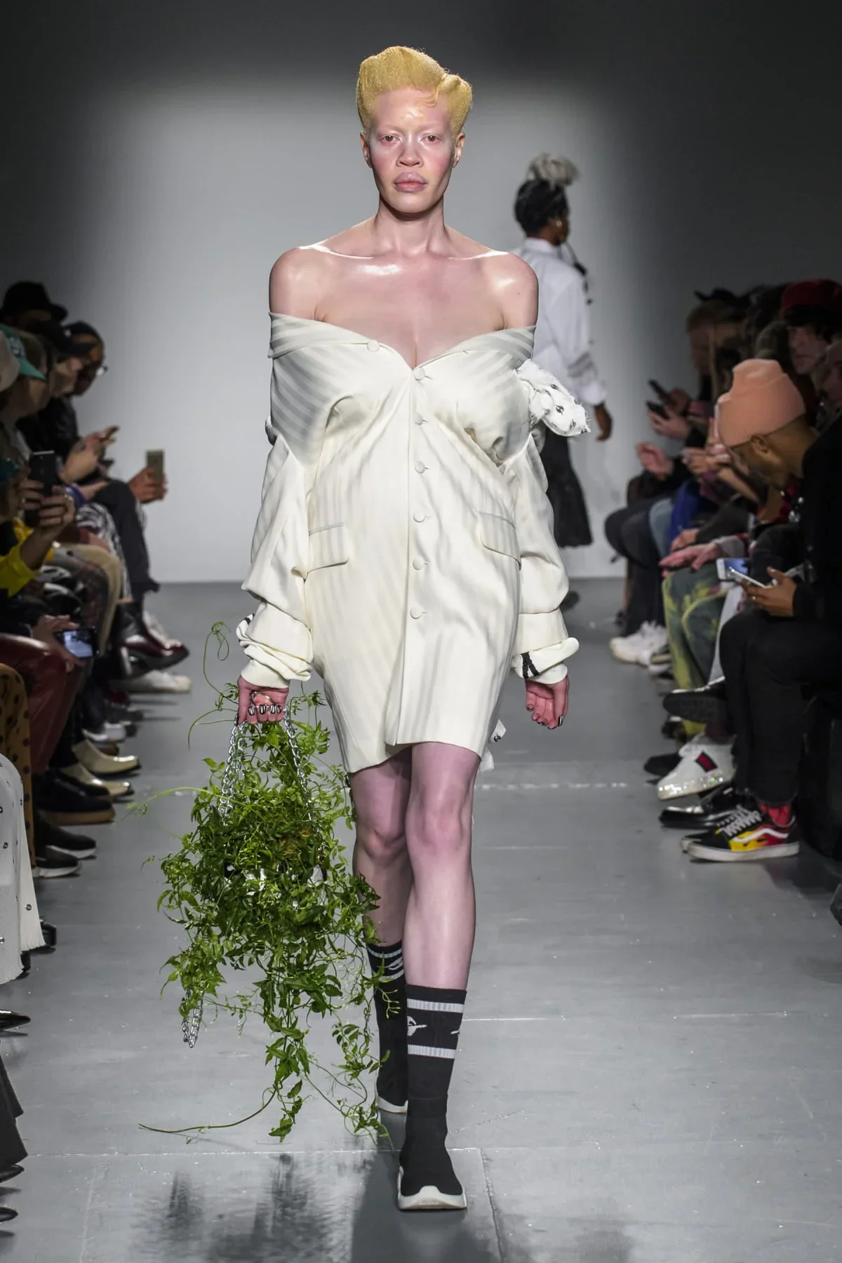 A model walking down a runway wearing an off-shoulder, oversized beige blazer dress with buttons, black and white striped socks, and holding a bunch of green leafy plants in her left hand. The model has short, platinum blonde hair and a pale complexion. The runway is flanked by seated audience members taking photos.