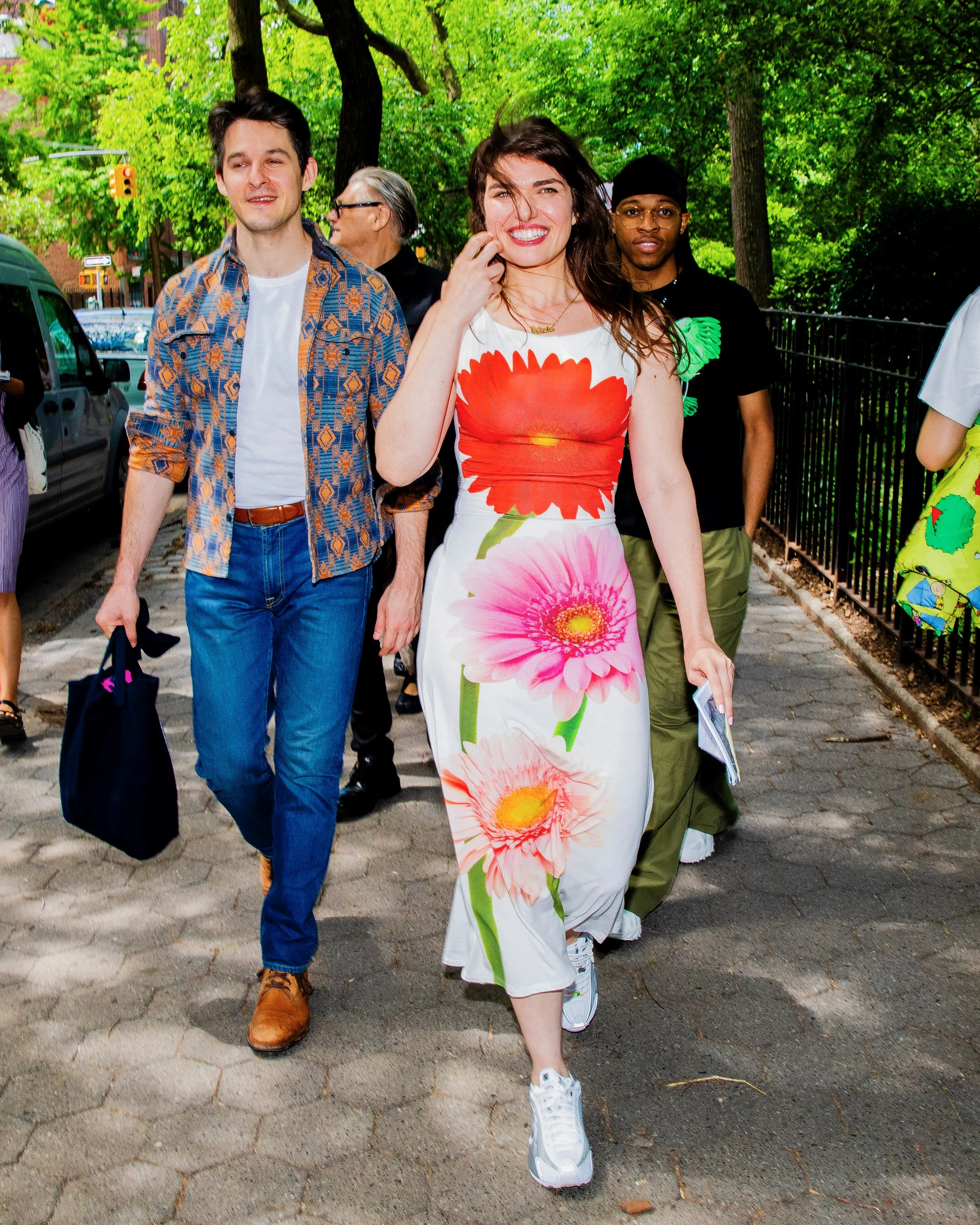 A woman in a white dress with large pink and red flower prints walking on a sidewalk, smiling, with several people in the background and green trees overhead.