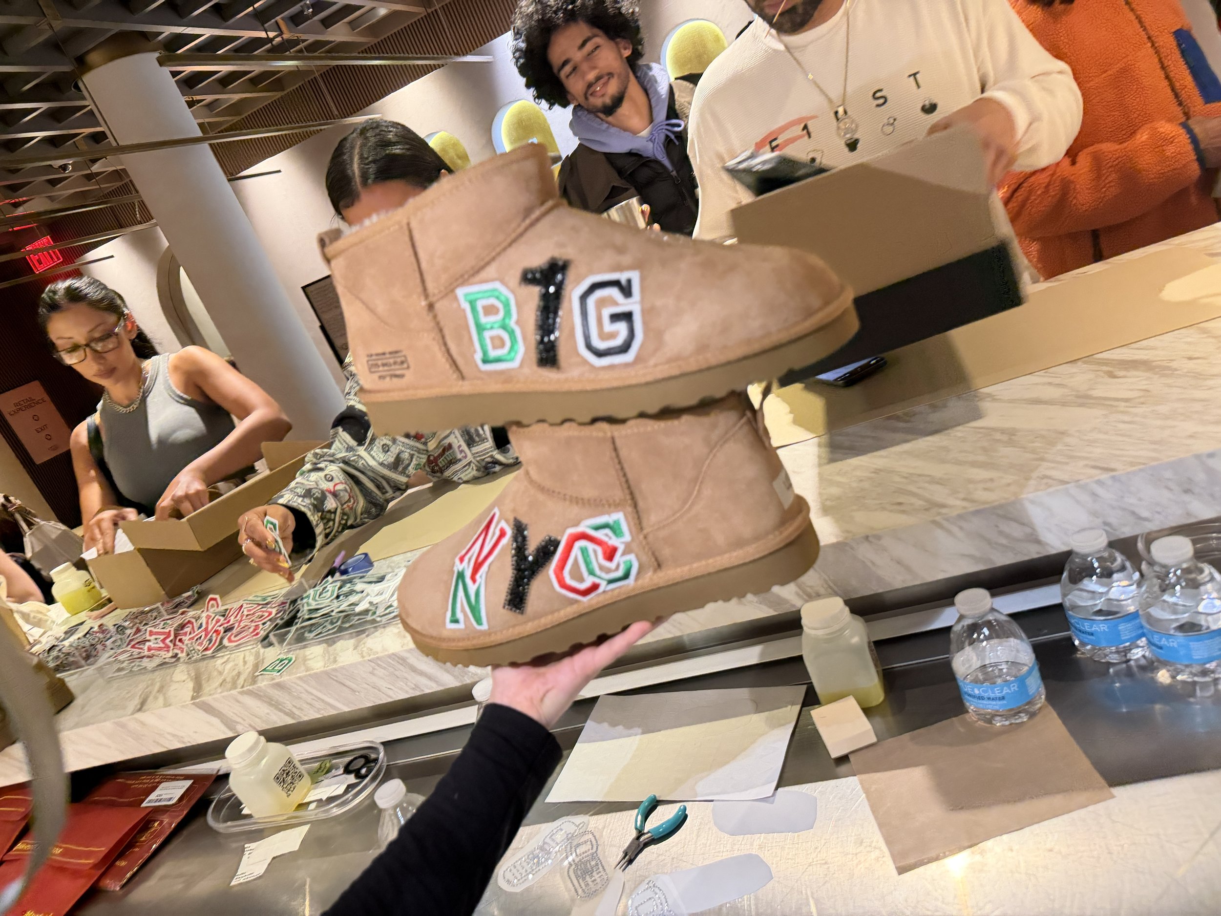 A tan pair of boots with decorative patches spelling 'BIG' and 'NYC' being held up in a store or decoration event