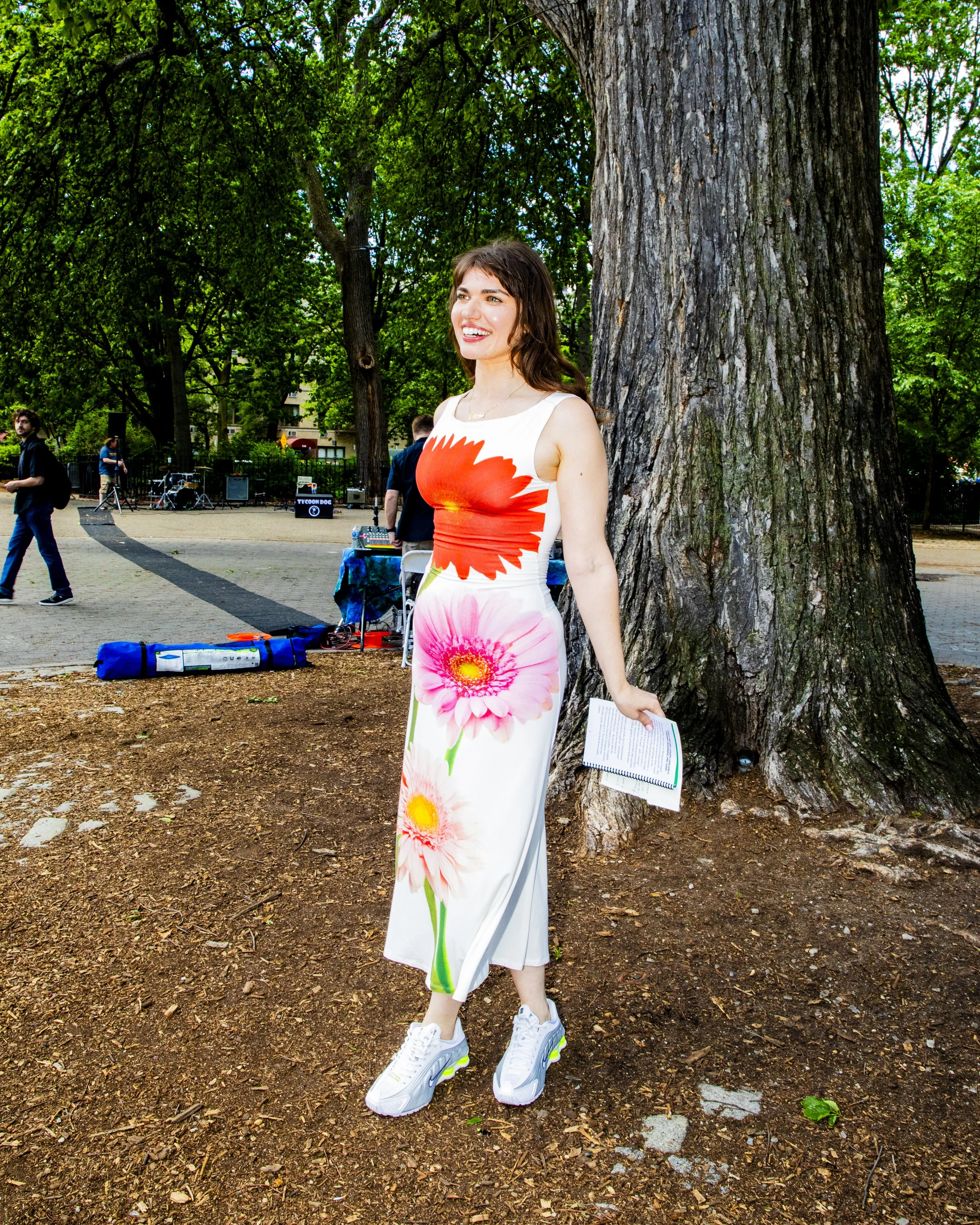 A woman standing outdoors next to a large tree, smiling, wearing a dress with large floral print, holding a notebook, in a park with people and tables in the background.