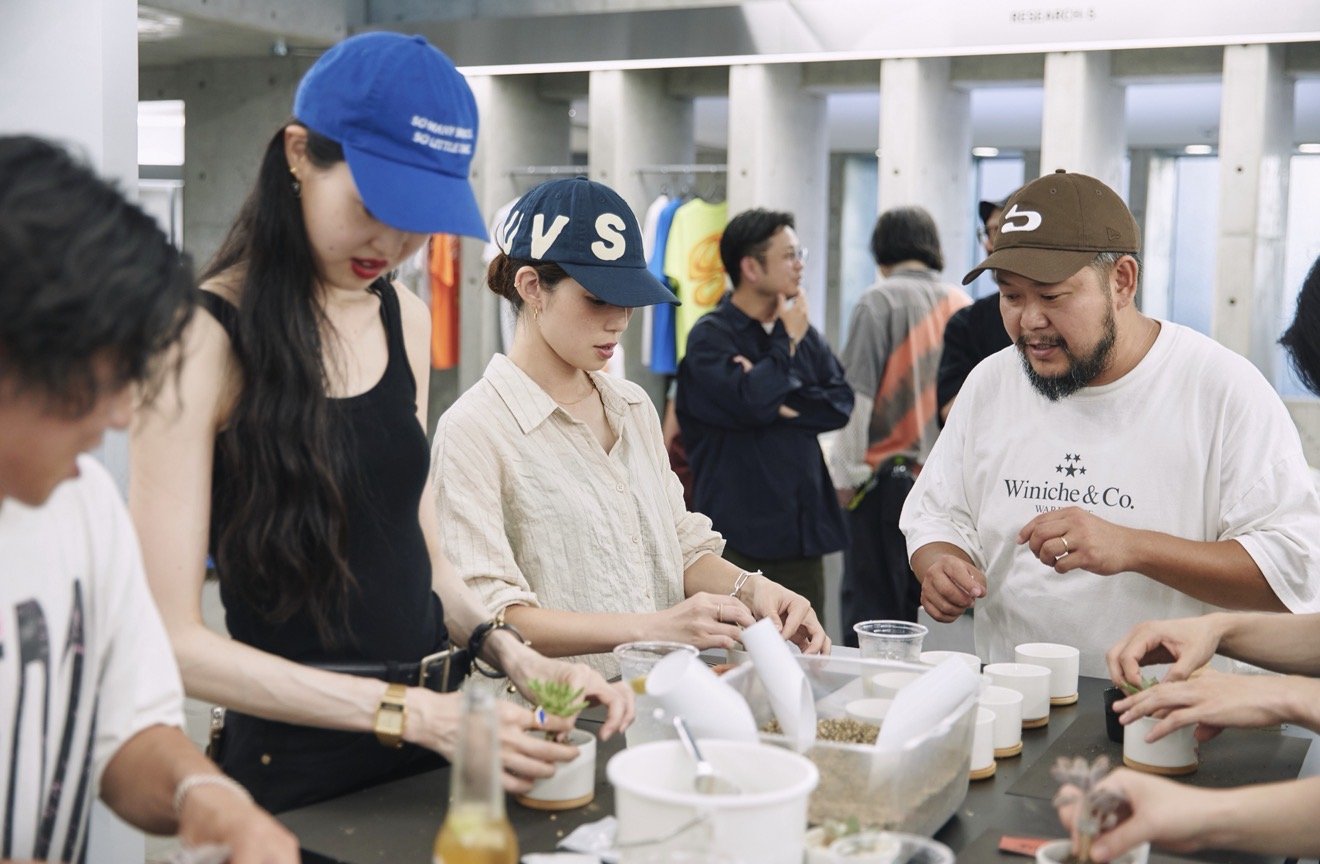 A group of people gathered around a table participating in a workshop or craft activity inside a modern building with concrete pillars and clothing display in the background.