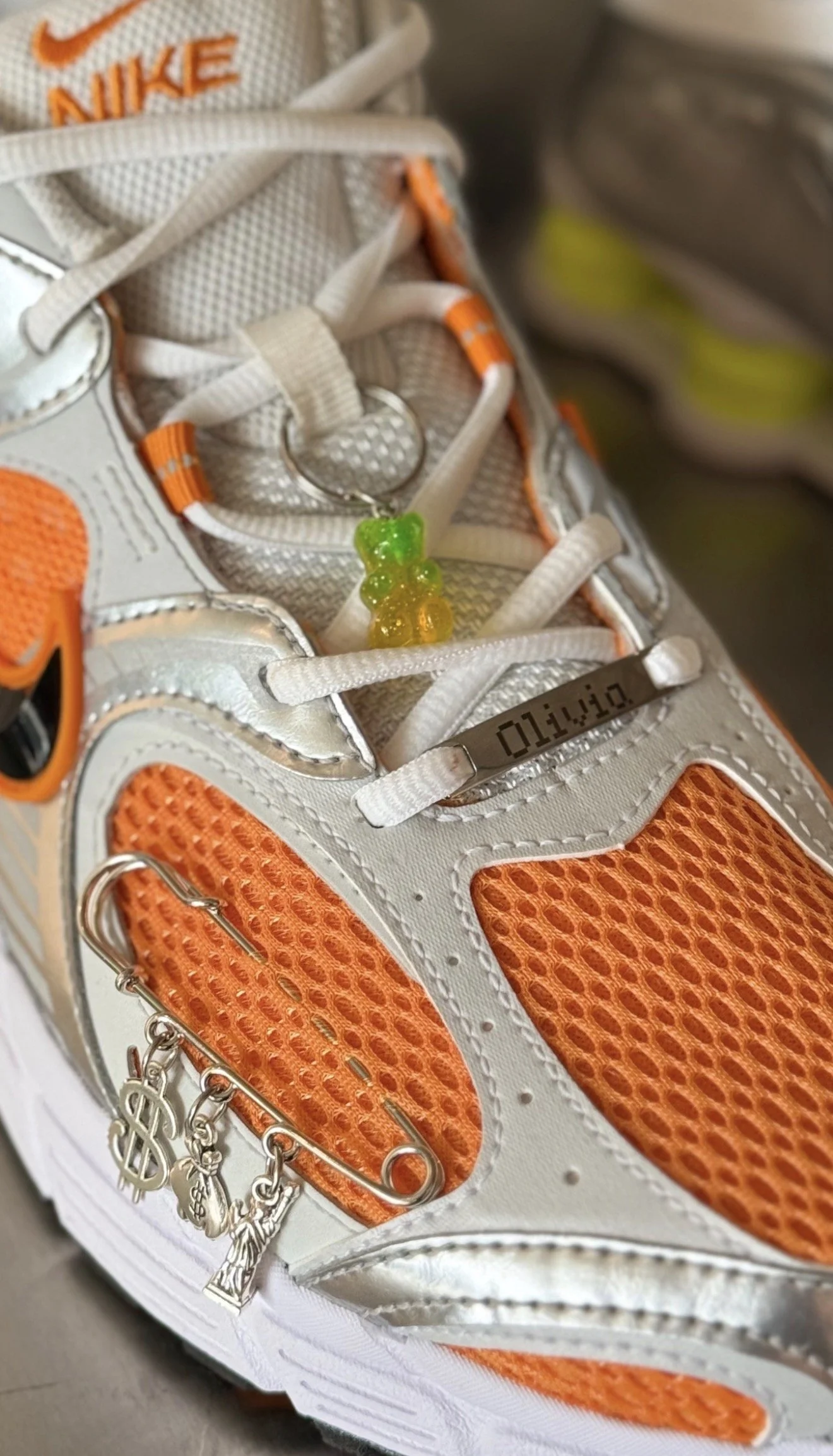 Close-up of a white and orange Nike athletic shoe with silver accents. Attached to the laces is a keychain with gummy bear charms and a digital ID tag reading 'OTAKU10'.