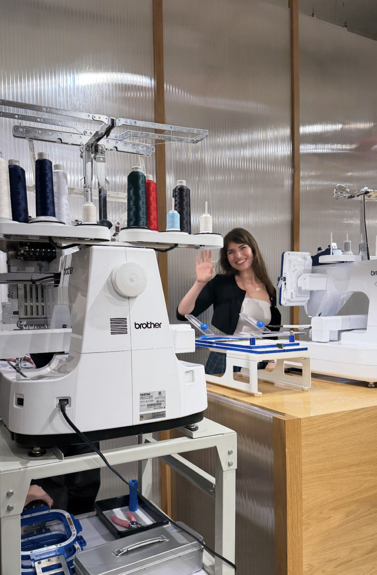 A woman smiling and waving at a sewing or embroidery workshop, surrounded by sewing machines, spools of thread, and sewing tools.