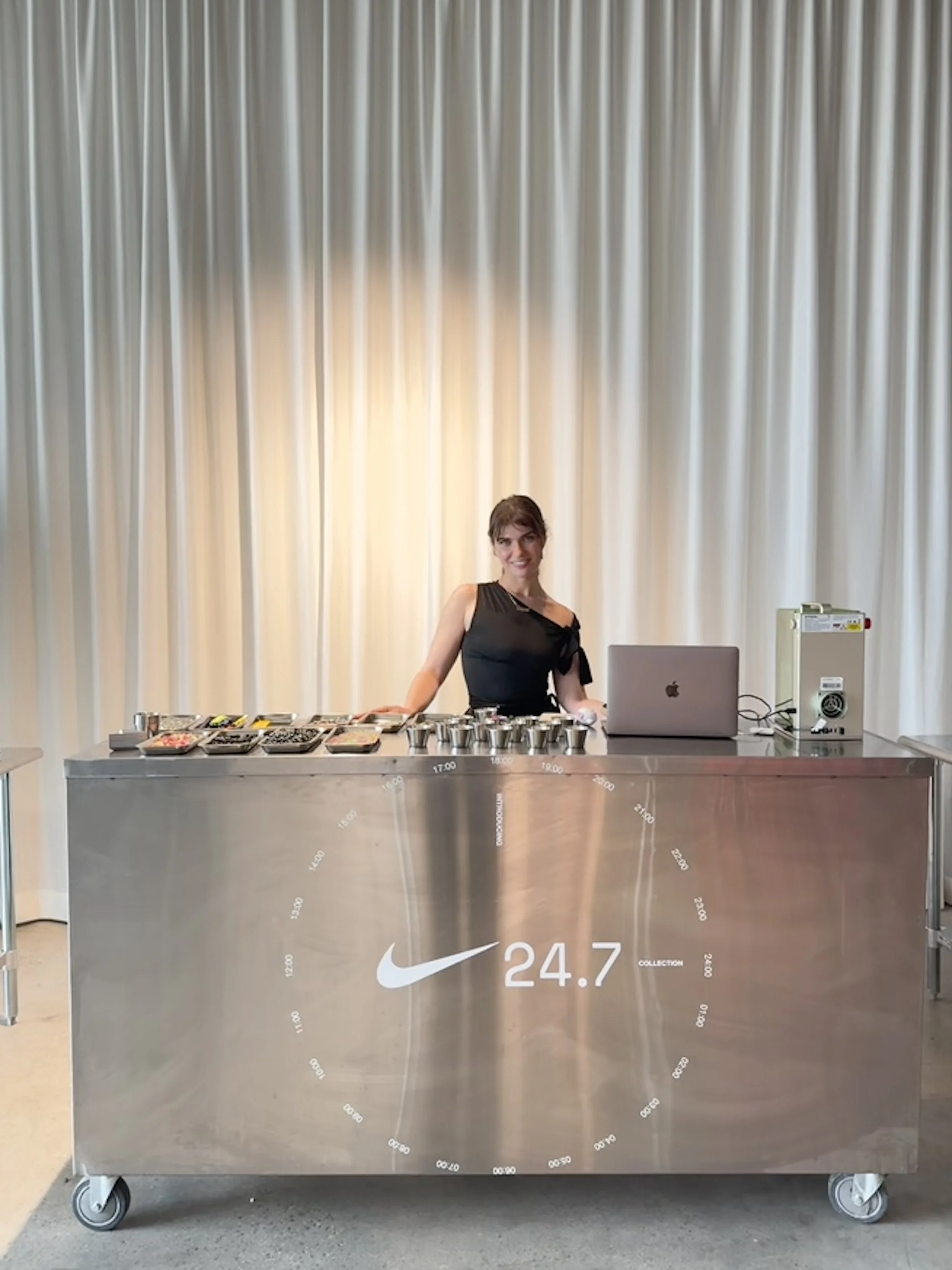 Woman standing behind a Nike-themed stainless steel table with various containers, a laptop, and electronic equipment, in front of a gold curtain.