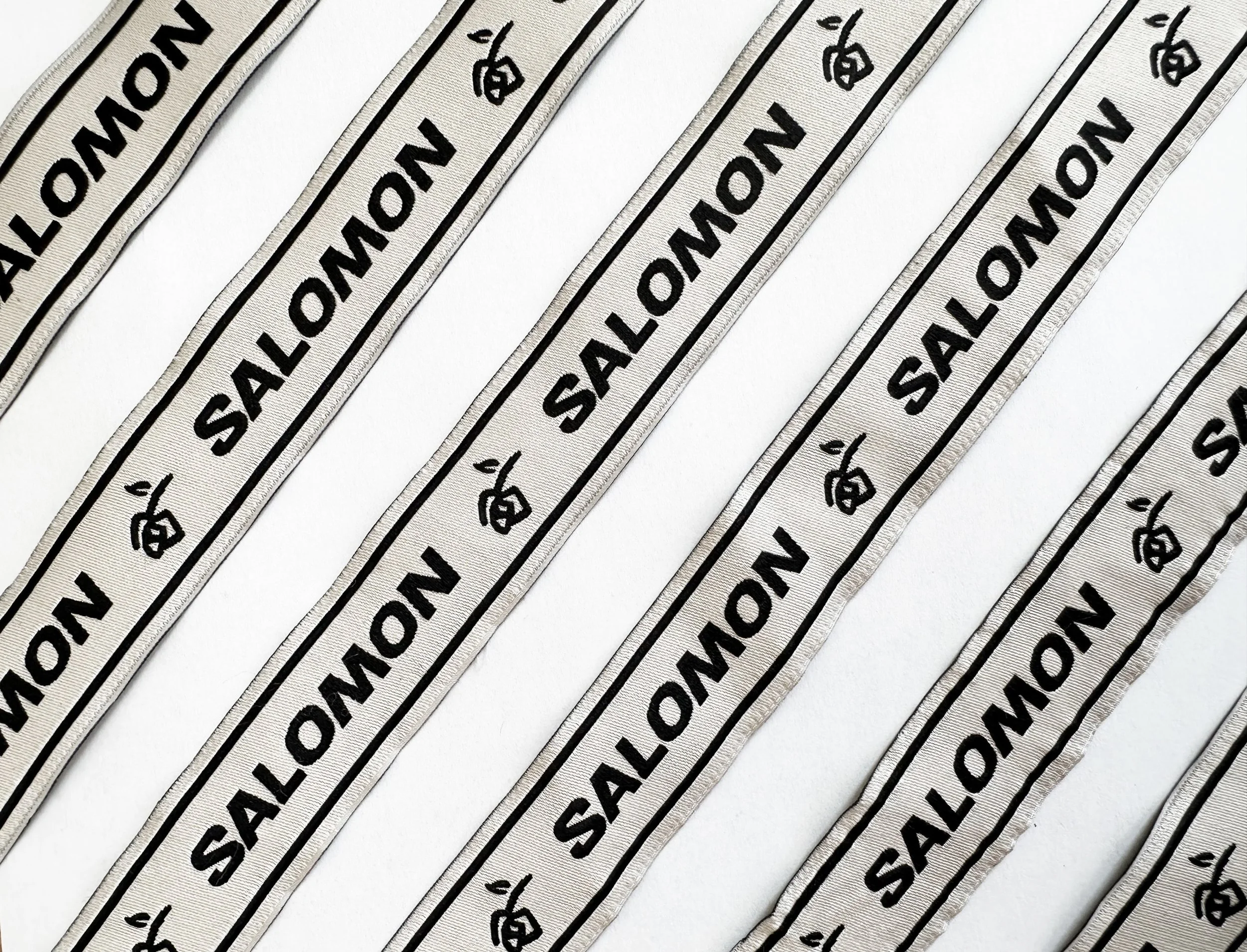 Strip of fabric labels with the text 'SALOW' in bold black letters, along with a small logo, repeated across multiple strips on a white background.
