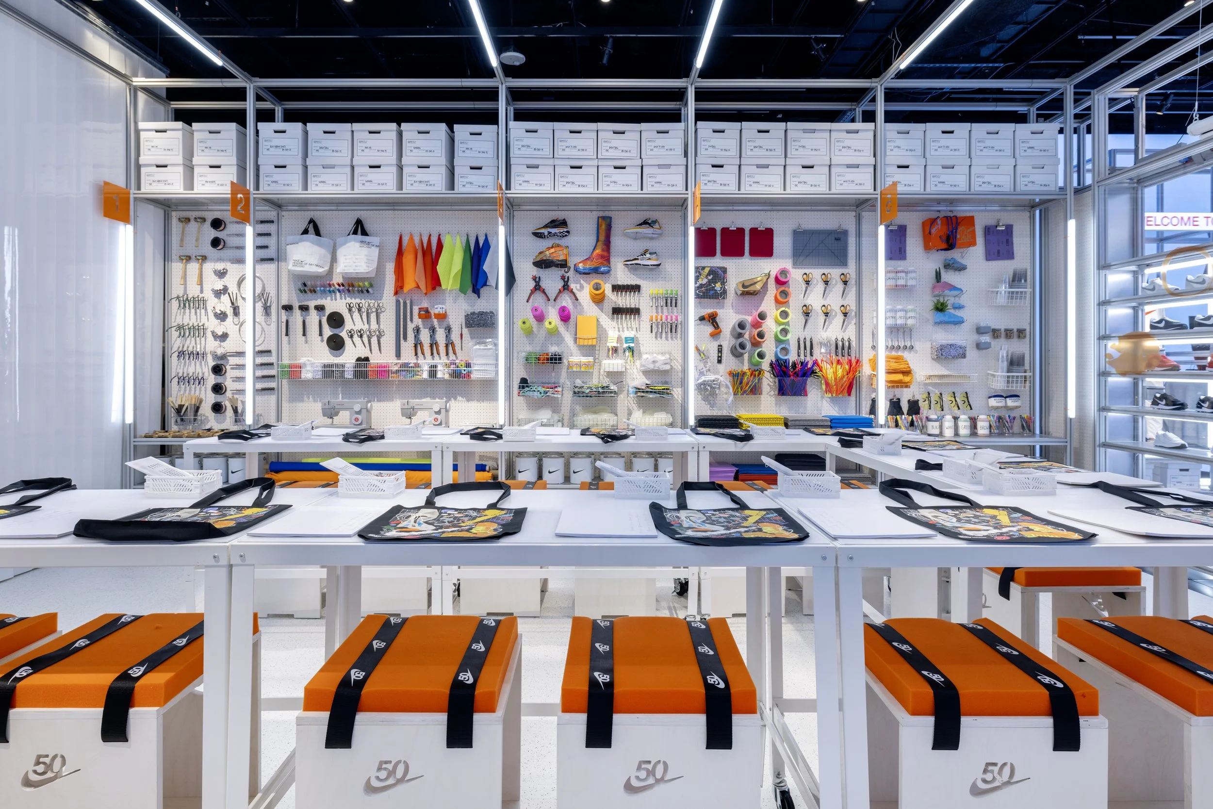 A tech store display with an organized wall of assorted office and sports supplies, including scissors, tape, paints, markers, and shoes, along with tables of orange and white stools and backpacks, illuminated with bright lighting.