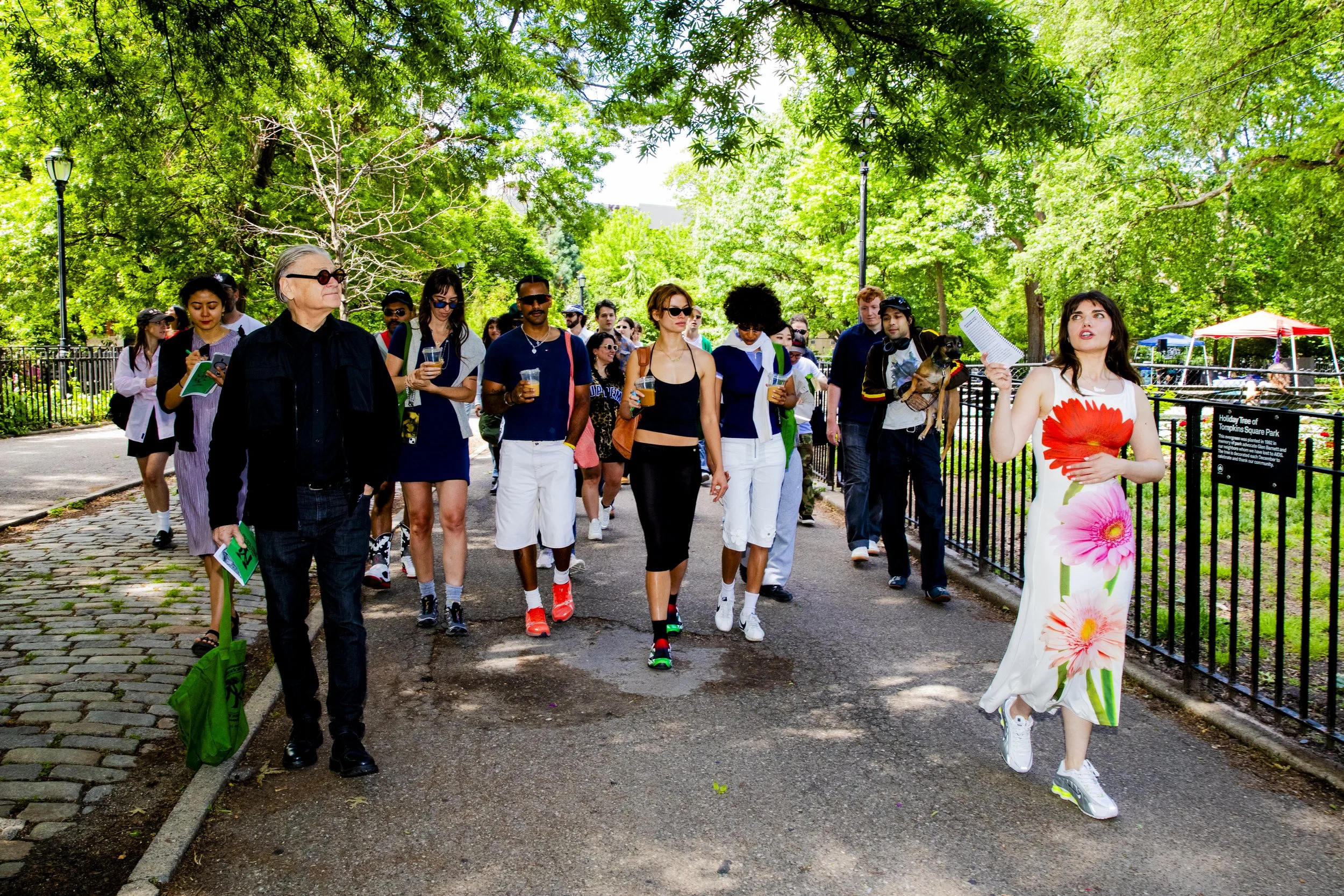 A group of people walking through a park, led by a woman in a white dress with large pink and red floral patterns, holding a paper, with trees and greenery around them.