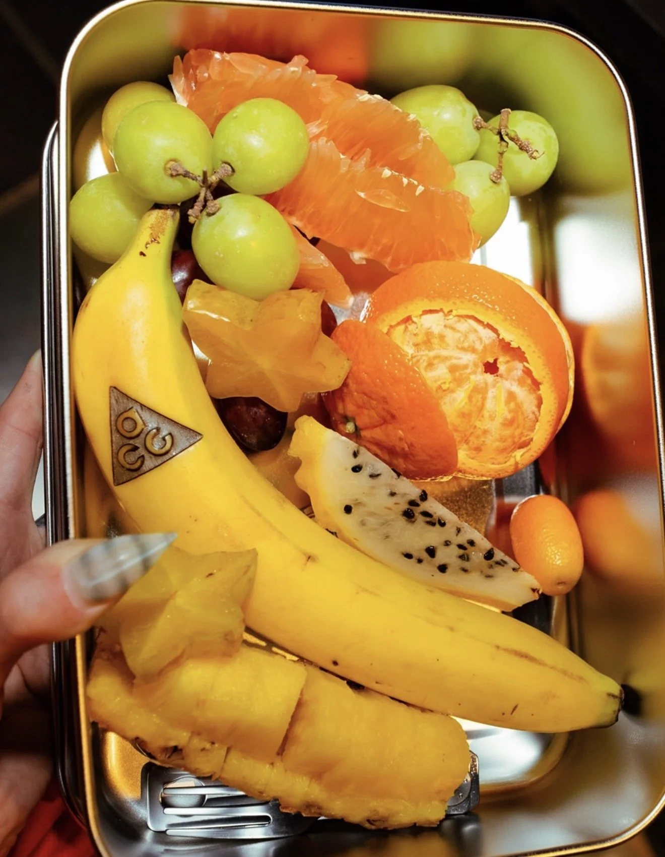 Assorted fresh fruits including green grapes, a banana, orange segments, pineapple chunks, dragon fruit slice, starfruit, a peeled orange, and kumquats in a metal tray.