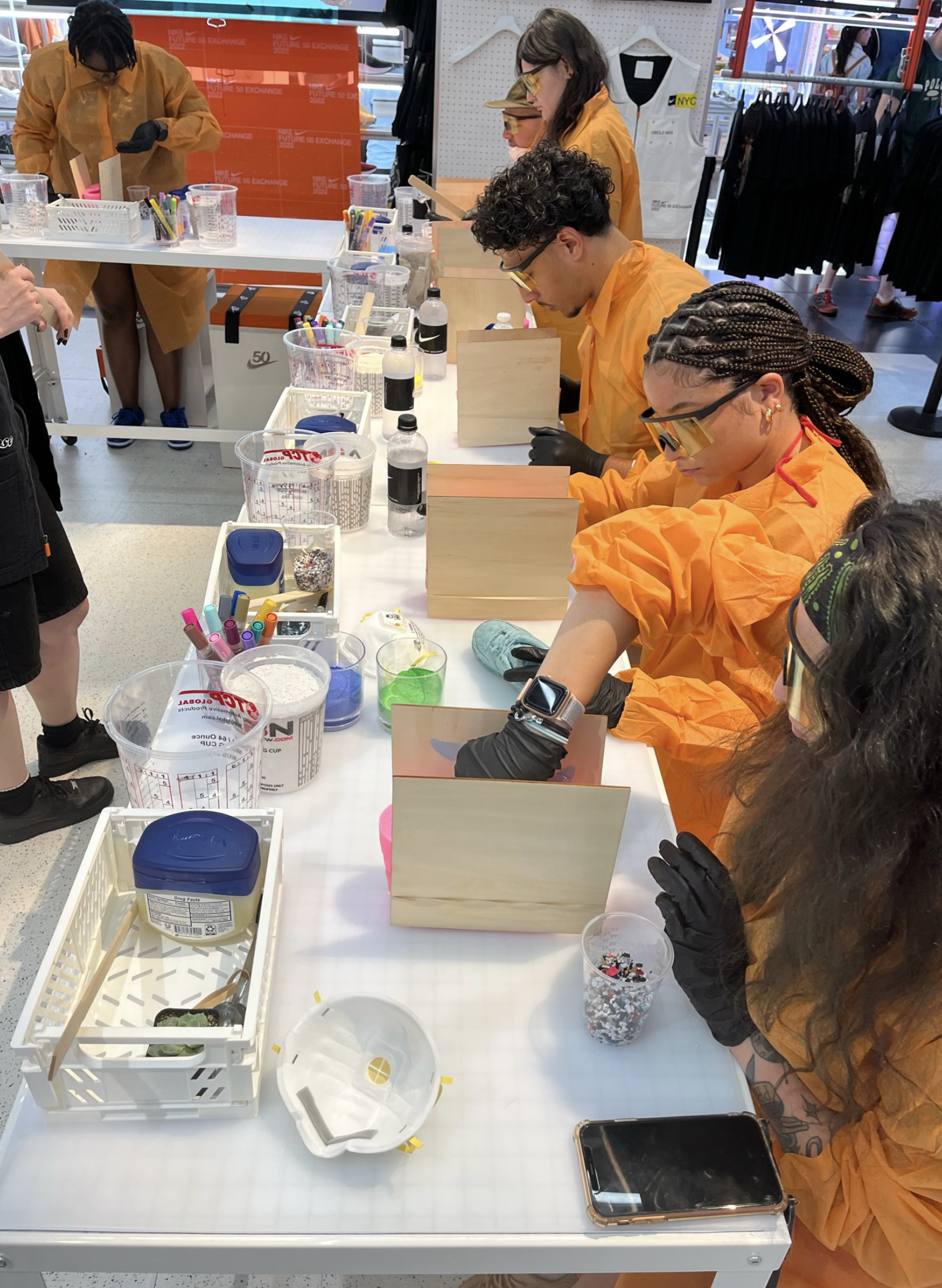 Group of people wearing orange protective smocks and black gloves engaging in a craft activity at a table inside a retail store.
