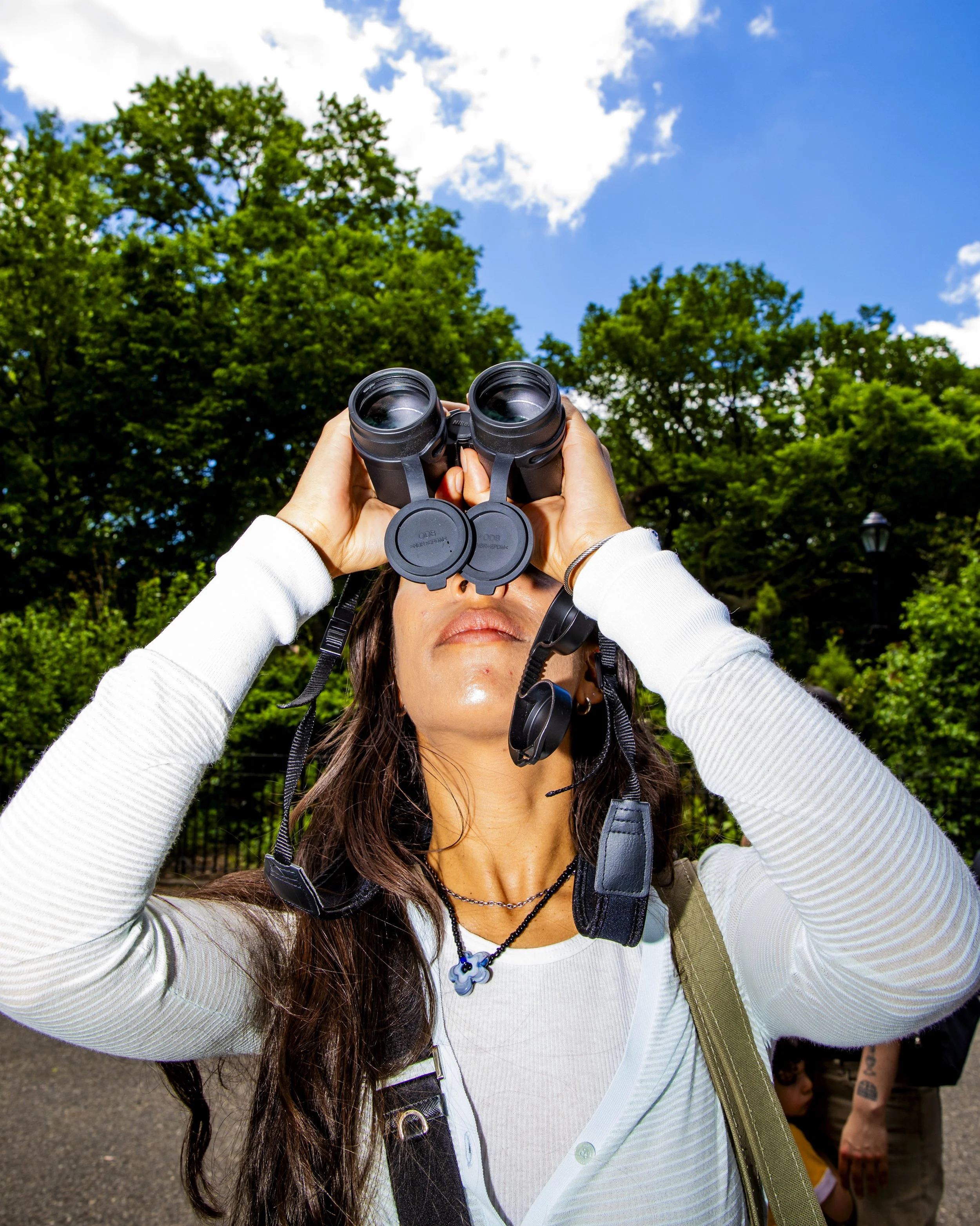 Woman looking through binoculars outdoors with trees and a blue sky in the background.