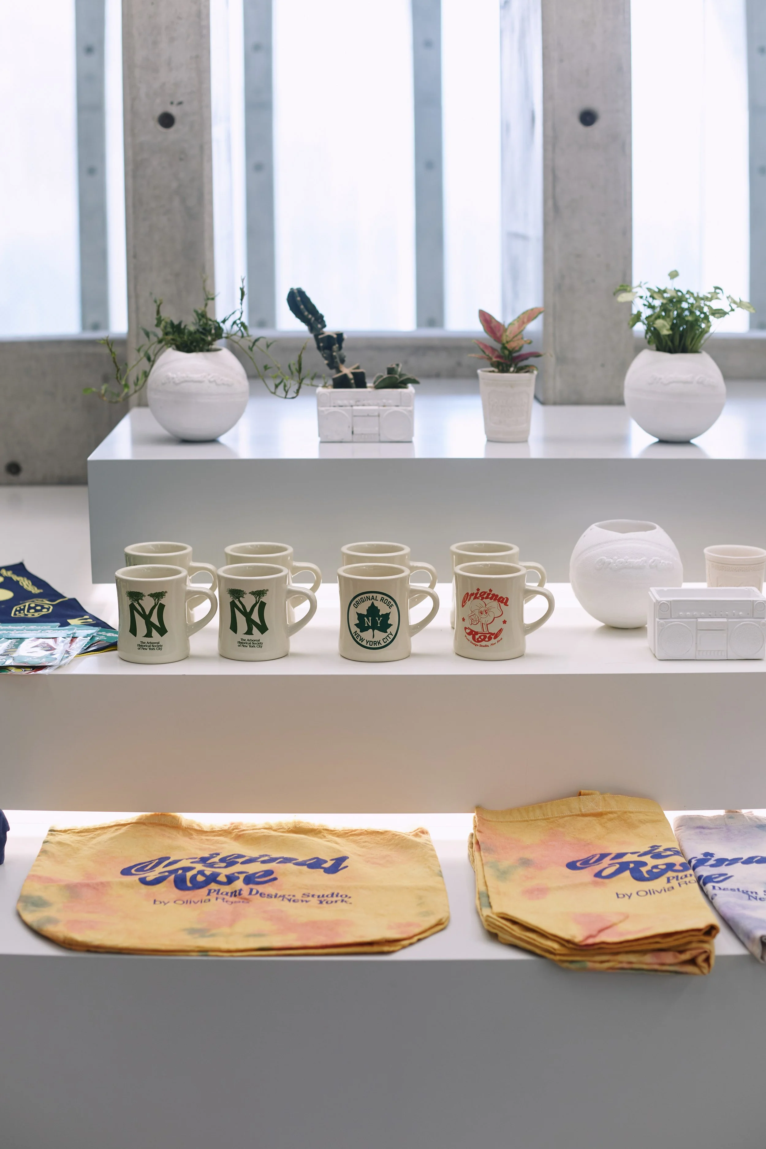 Display of souvenir items on a white table, including ceramic mugs with New York City logos, tie-dye cloth bags with "Odeon Rose" branding, small potted plants, and a vintage radio.