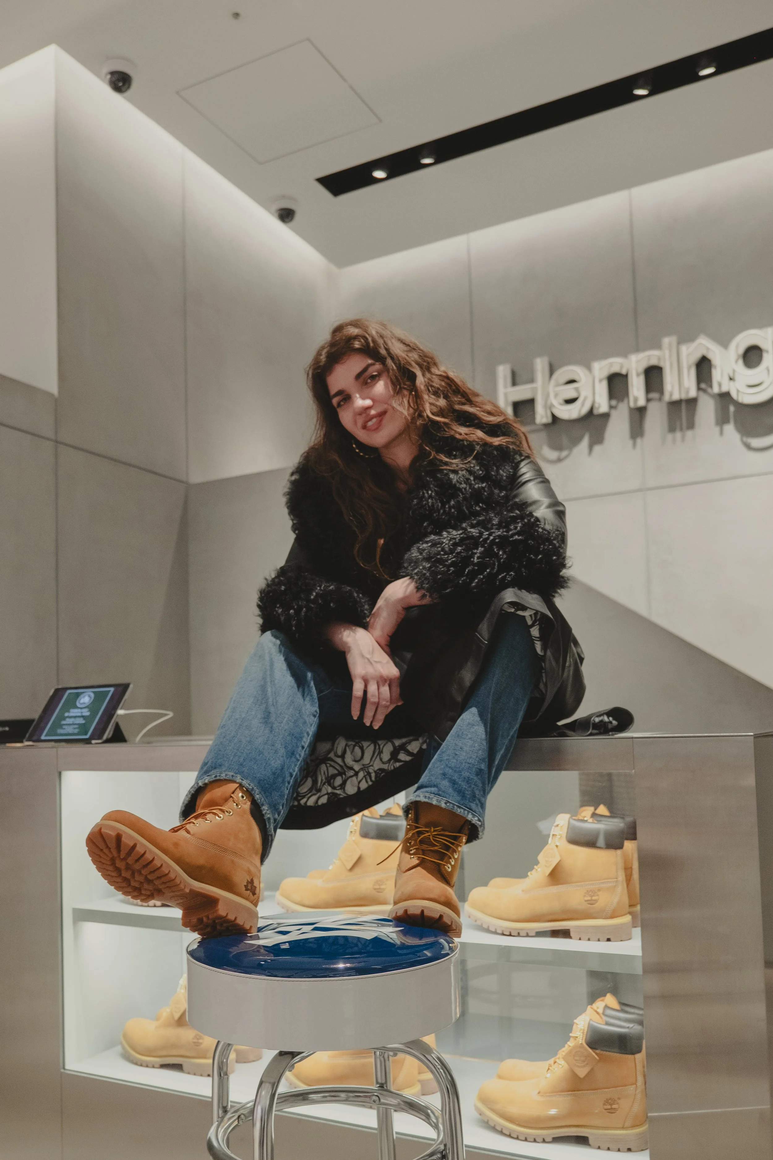 A young woman with wavy brown hair wearing a black faux fur jacket, blue jeans, and tan Timberland boots sitting on a display counter inside a shoe store, with Timberland shoes in the background and a sign that says "Herring" on the wall.