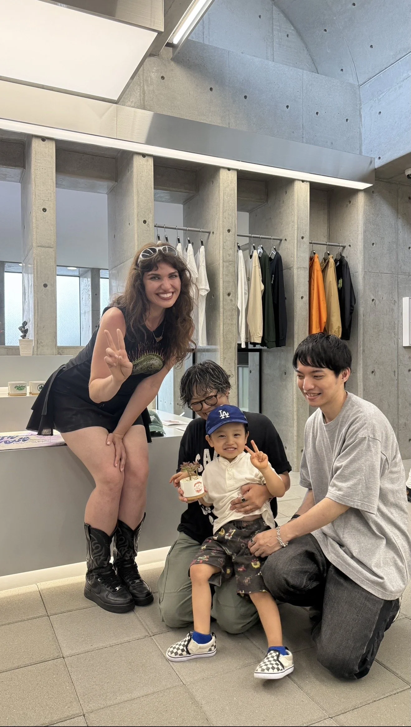 Four people, including a boy holding a plant, are posing and smiling indoors, with clothing hanging on the wall behind them.