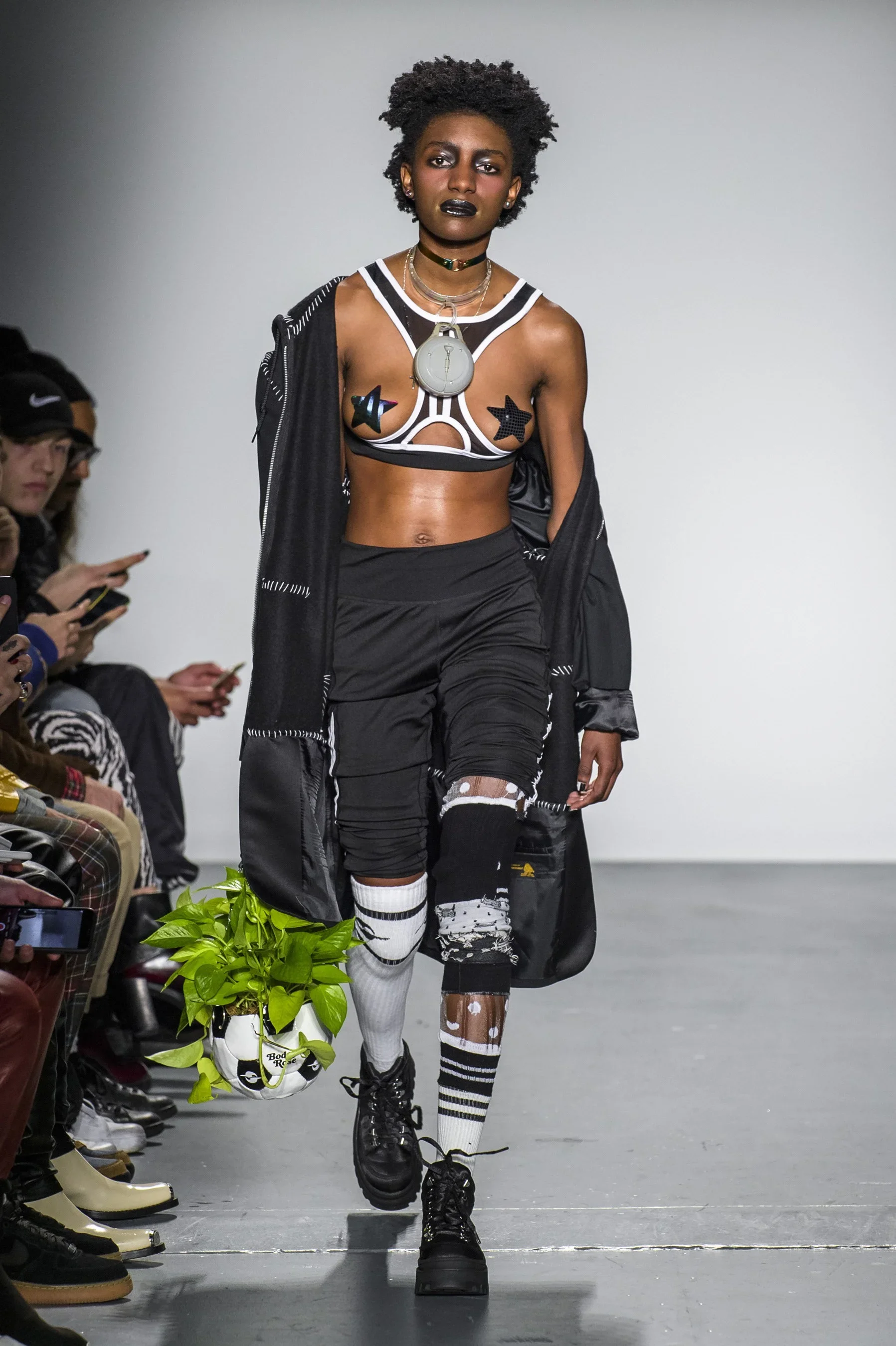 A fashion model walks down a runway wearing a black and white athletic crop top, black distressed pants with ripped knee, black combat boots, and mismatched knee-high socks, one white with black stripes and the other brown with white dots. She carrie