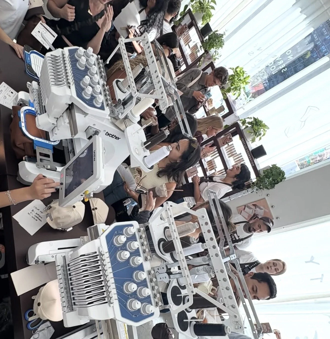 People gathered around a table at a sewing or embroidery workshop, with sewing machines and fabric samples, in a well-lit room with large windows and potted plants.