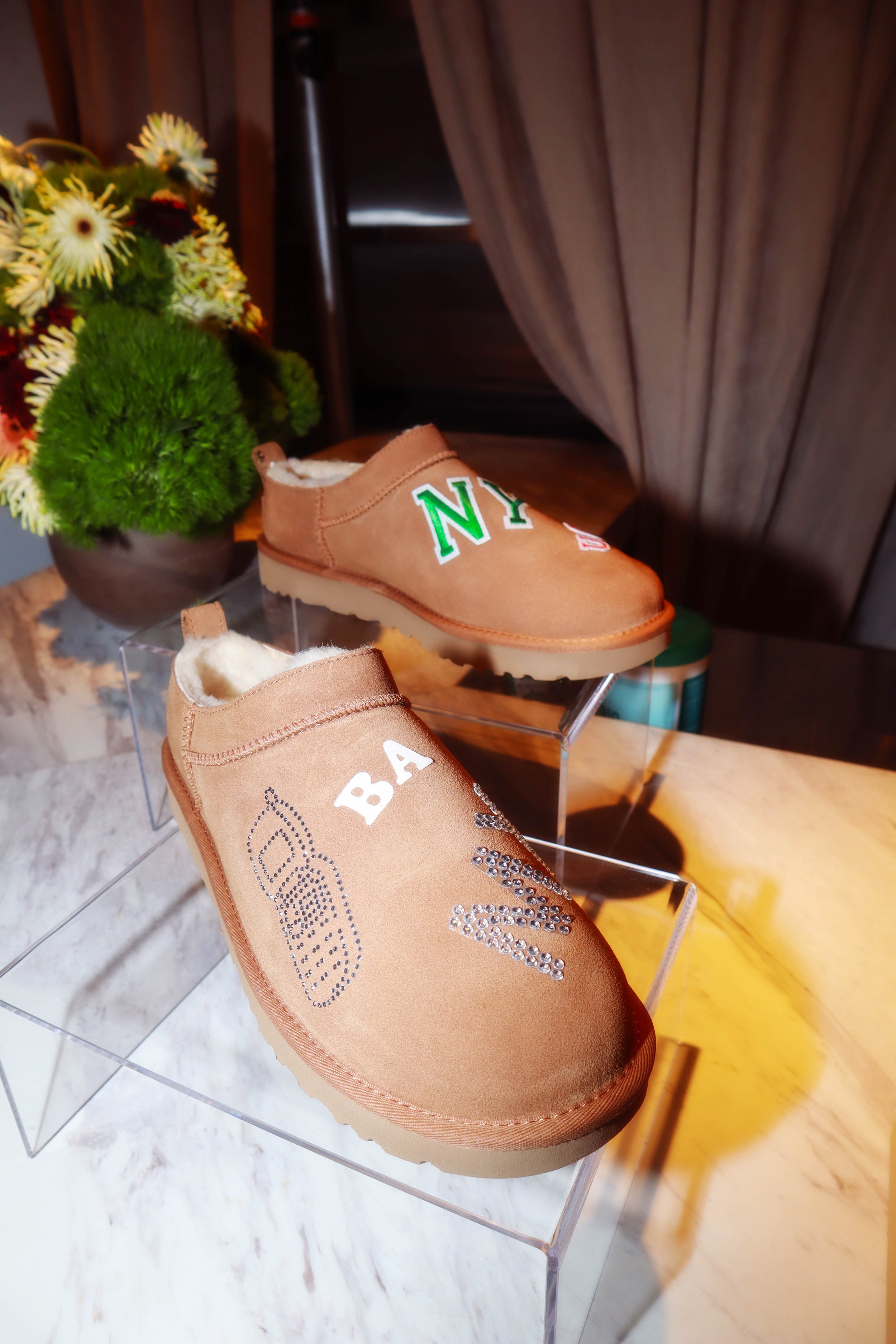 Two tan slip-on shoes with decorative rhinestones and embroidered text, one with 'BABI' and the other with 'NY', displayed on clear stands with a flower arrangement and brown curtains in the background.