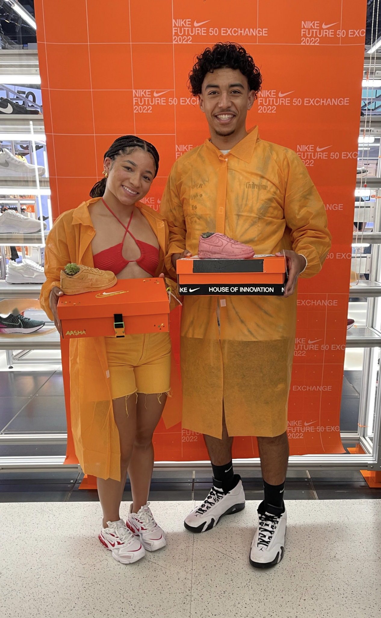 Two individuals standing in front of a Nike event backdrop, holding shoeboxes with sneakers on top. The woman on the left wears a red crop top and yellow shorts, and the man on the right wears a yellow coat and tie-dye shorts. Both are smiling, and t