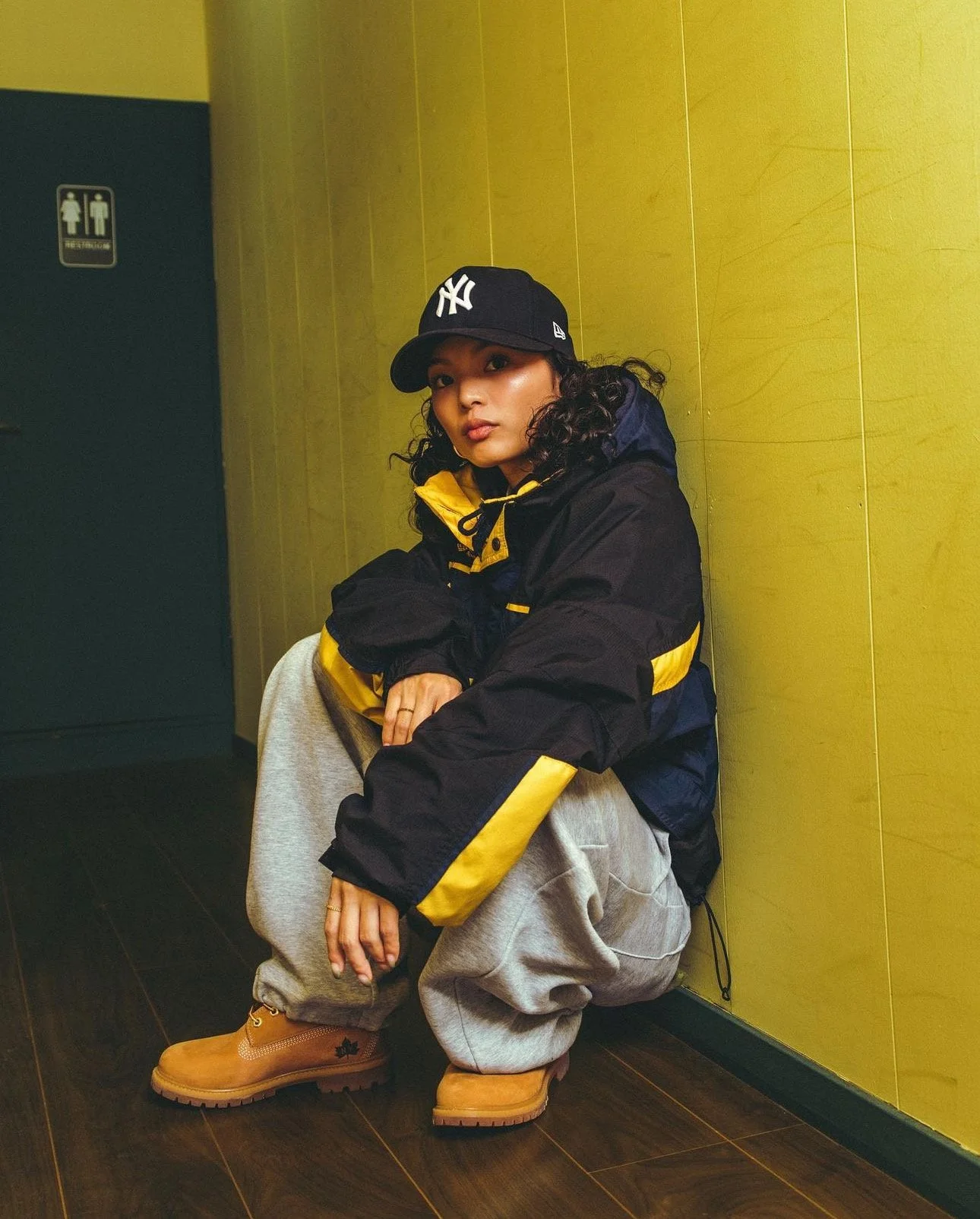 A young woman wearing a black and yellow jacket, gray sweatpants, tan Timberland boots, and a black baseball cap squats against a yellow wall in a room with dark wood flooring.