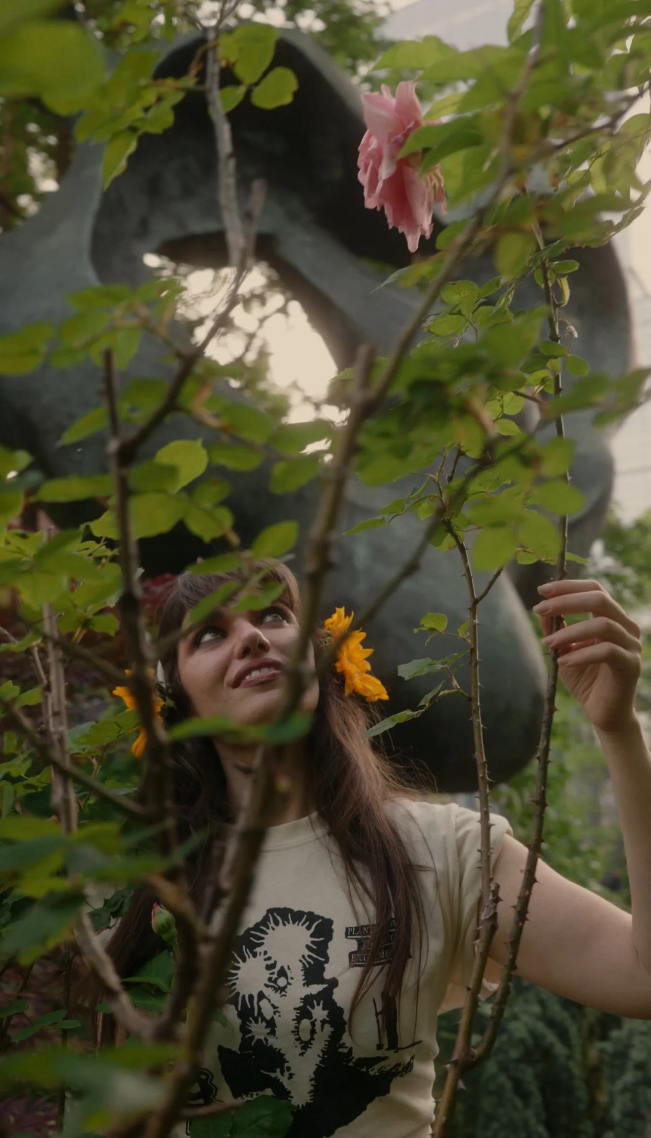 A young woman with long brown hair wearing a beige t-shirt with a black graphic design, surrounded by greenery, is looking up through leaves and branches, with pink and yellow flowers in her hair, and a large sculpture in the background.