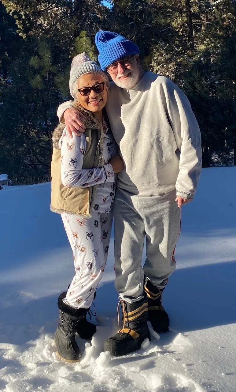 Two elderly people standing in snow outdoors during winter, smiling at the camera. They are dressed in warm clothing, with one person wearing a blue knit hat and the other wearing a gray knit hat.