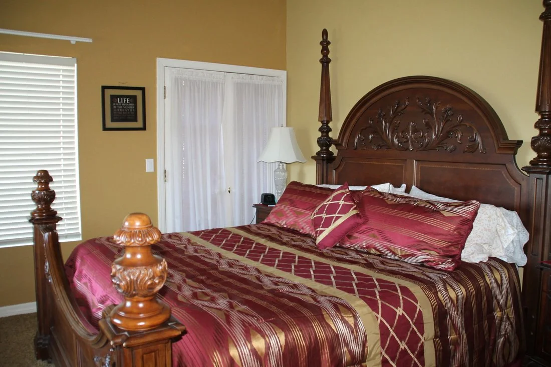 A bedroom with a large wooden bed featuring an ornate headboard and posts, red and gold striped bedding, pillows, a nightstand with a lamp, a window with white blinds, and a wall with a framed picture.