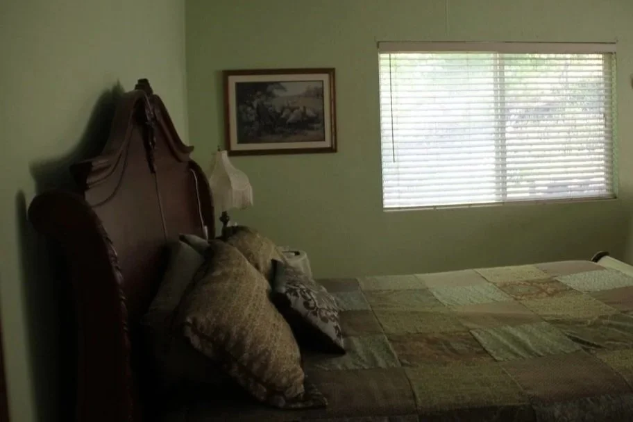 A bedroom with a large bed, multiple pillows, a wooden headboard, a window with blinds, and a framed landscape picture on a light green wall.