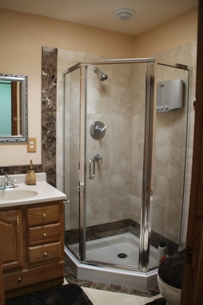 Bathroom with a glass shower enclosure, a wooden vanity with a mirror, and a toilet. The shower has a handheld showerhead and a soap dispenser. The walls are tiled in beige with dark accents.
