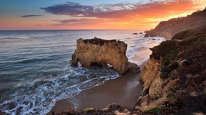 Sunset over a coastal scene with a natural rock arch, sandy beach, and cliffs with vegetation.
