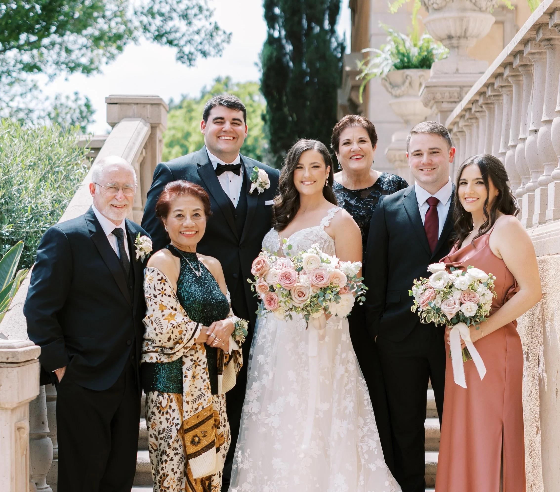 Group of people at a wedding, including bride in white dress holding a bouquet, groom in black tuxedo, and family members dressed formally, standing on outdoor stone staircase with greenery and ornate stone planters in background.
