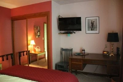 A hotel room with a bed, a wooden desk, a chair, and a wall-mounted TV. There is a framed picture on the wall and a lamp on the desk. An open doorway reveals a lit table and mirror in the next room.