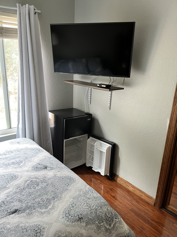 Room corner with wall-mounted TV, small shelf, mini fridge, and mini fridge door open, next to a bed with a patterned quilt, a window with curtains.