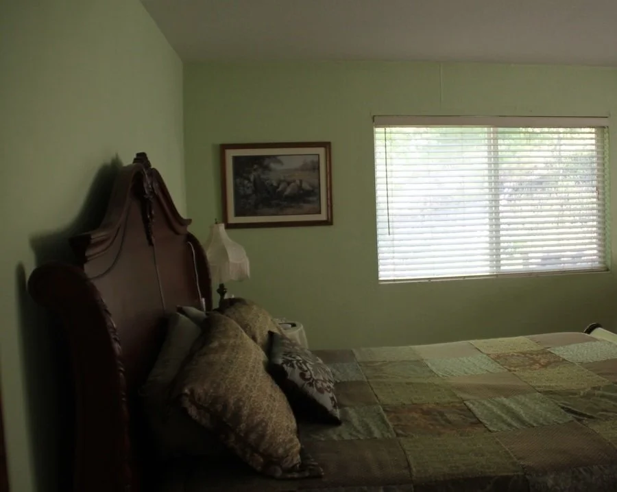 A bedroom with a green wall, a large window with blinds, a bed with patterned pillows and a patchwork quilt, a wooden headboard, a side lamp, and a framed picture on the wall.