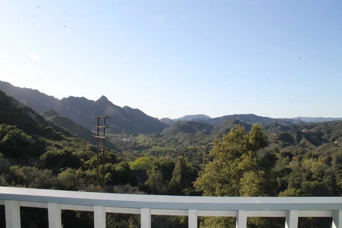 View of mountains and trees under a clear blue sky, with a utility pole in the foreground and part of a white railing at the bottom of the image.