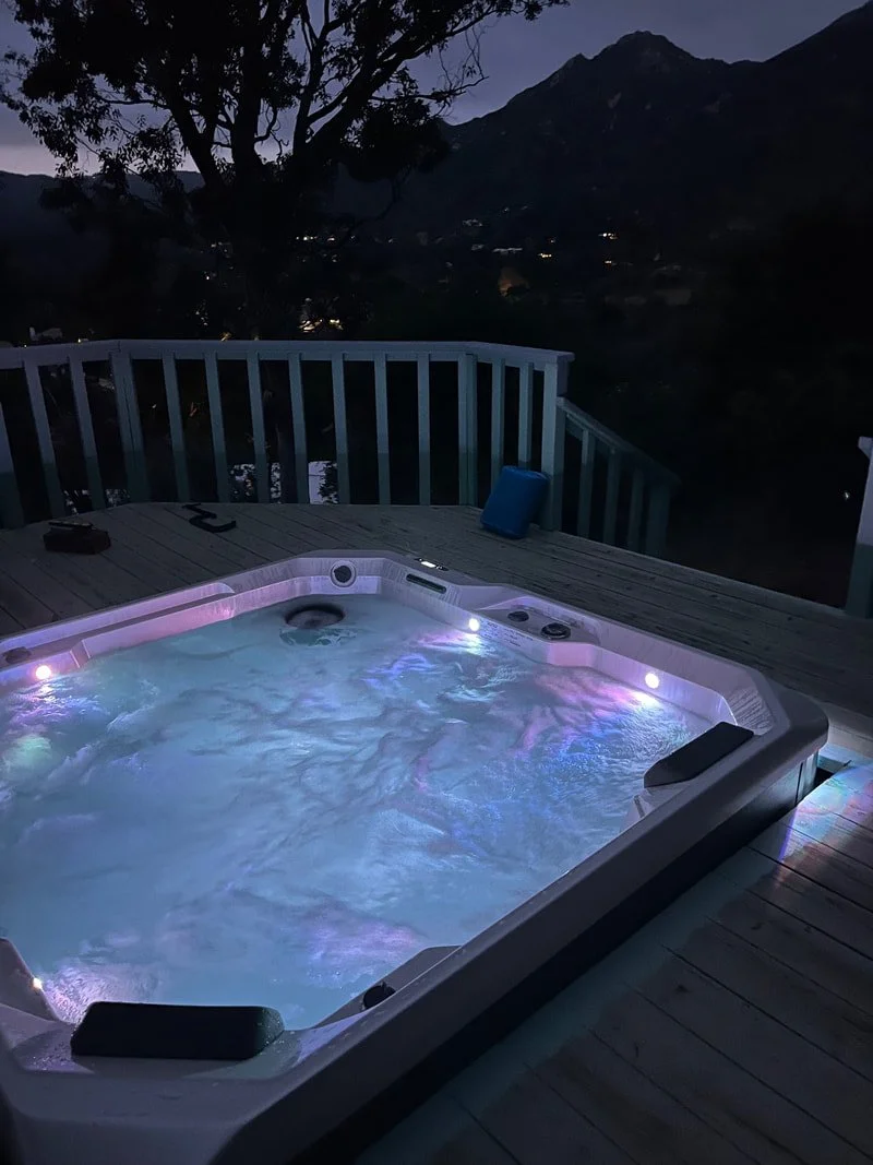 Nighttime scene showing a hot tub on a wooden deck, with a tree and mountains in the background.