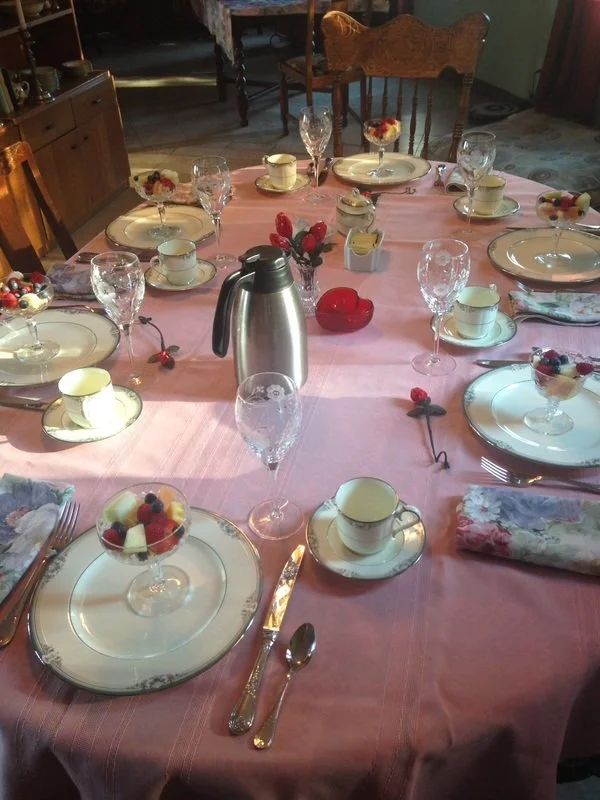 A family table setting with pink tablecloth, floral-patterned napkins, and fine china. The table has glass dessert bowls with fruit and cheese, cups and saucers, water glasses, a stainless steel pitcher, and a small red heart-shaped box. There are decorative cherry stems on the table, and a wooden chair is visible in the background.