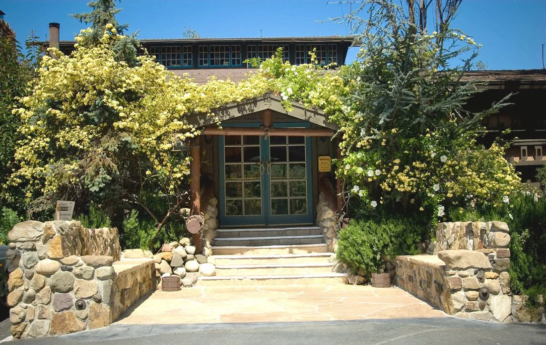 Front entrance of a house with a stone stairway, flanked by stone walls, surrounded by large flowering bushes, and topped with a wooden awning, with a clear blue sky overhead.