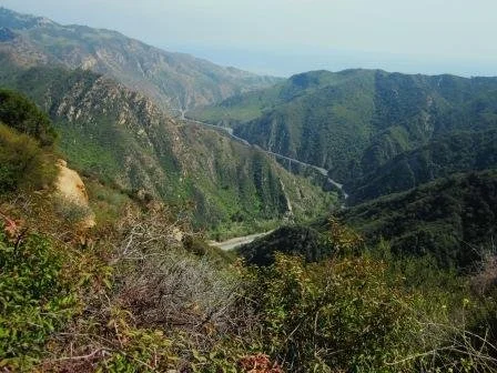 Mountain landscape view with green hills, a winding road, and a bridge in the distance.