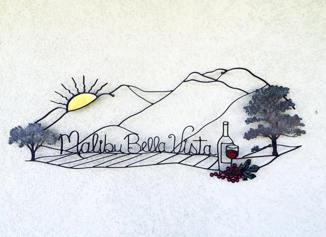 Metal wall art featuring mountains, sun, trees, a wine bottle, a glass of red wine, grapes, and the text 'Malibu Bella Vista'.