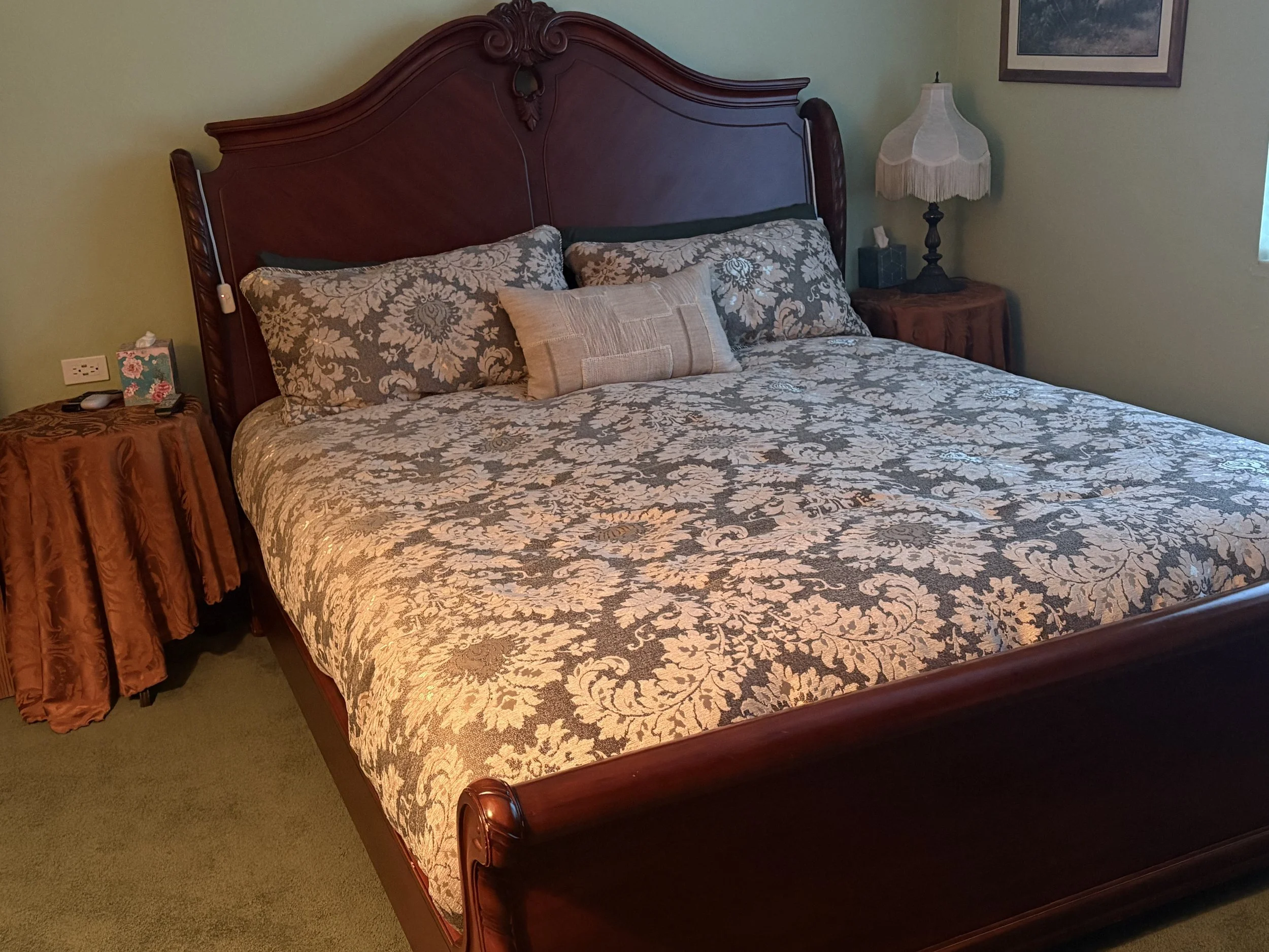 A large wooden bed with floral bedding and multiple pillows, situated in a cozy bedroom with side tables, lamps, and framed artwork on the wall.