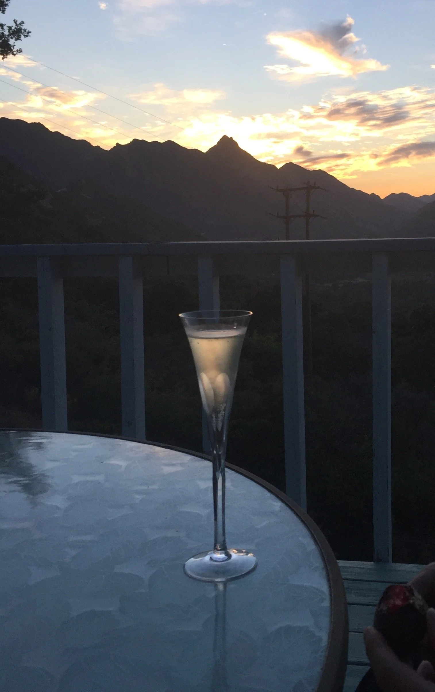 A tall champagne glass filled with a sparkling beverage on a glass table, with a sunset over mountain range in the background and power lines, on a balcony or patio.