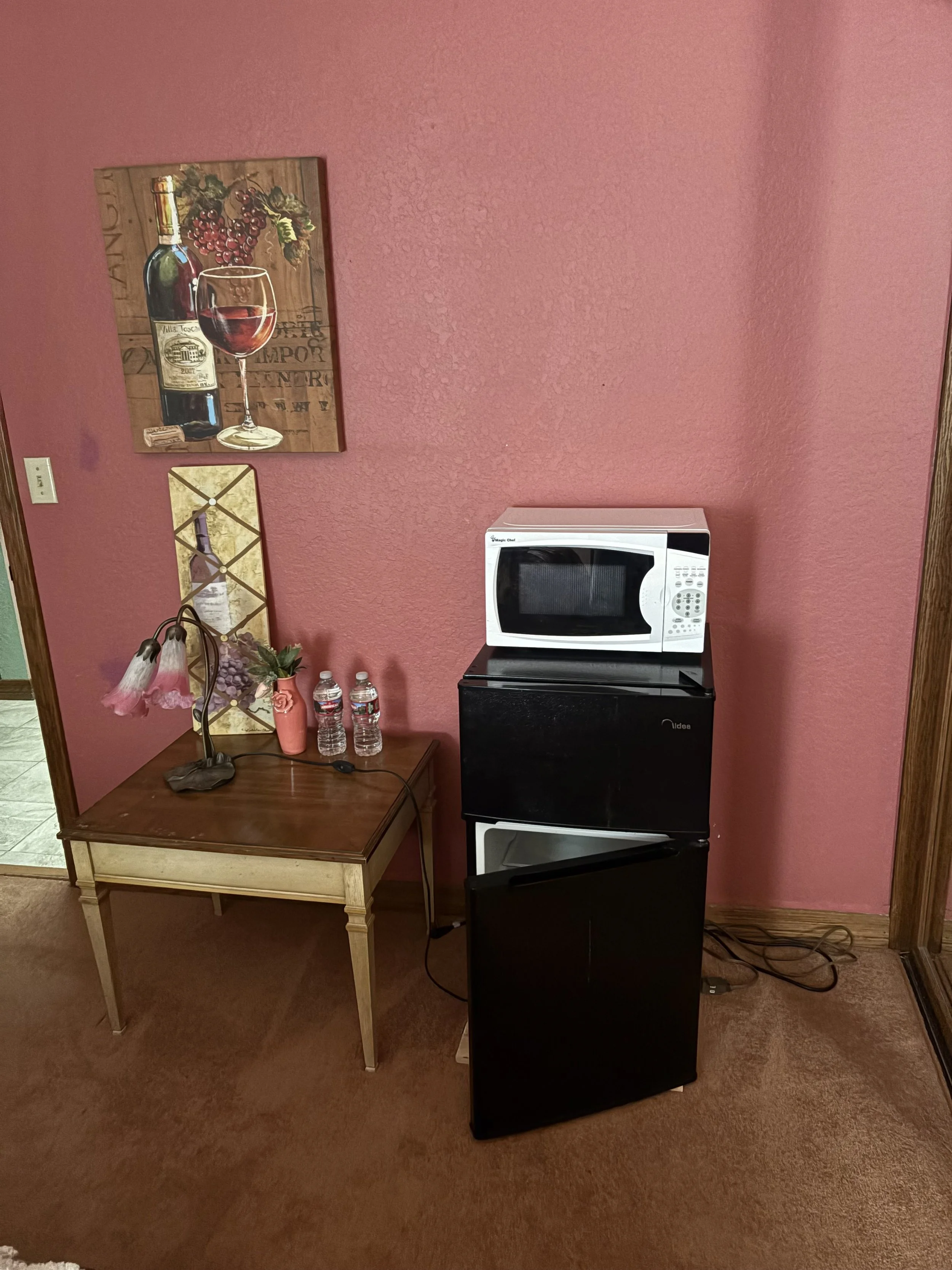Pink wall with wall art, a dark wood side table with a lamp, water bottles, and a vase, and a microwave on top of a small refrigerator.
