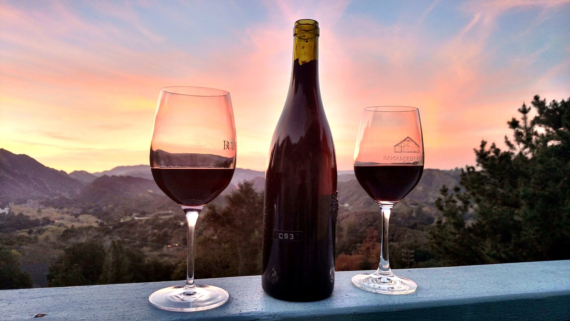 A wine bottle and two glasses of red wine on a table, with a sunset over mountainous landscape in the background.