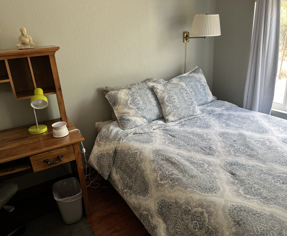 A neatly made bed with patterned duvet and pillows, beside a window with curtains, in a cozy bedroom with a wooden desk, green desk lamp, and wall-mounted lamp.