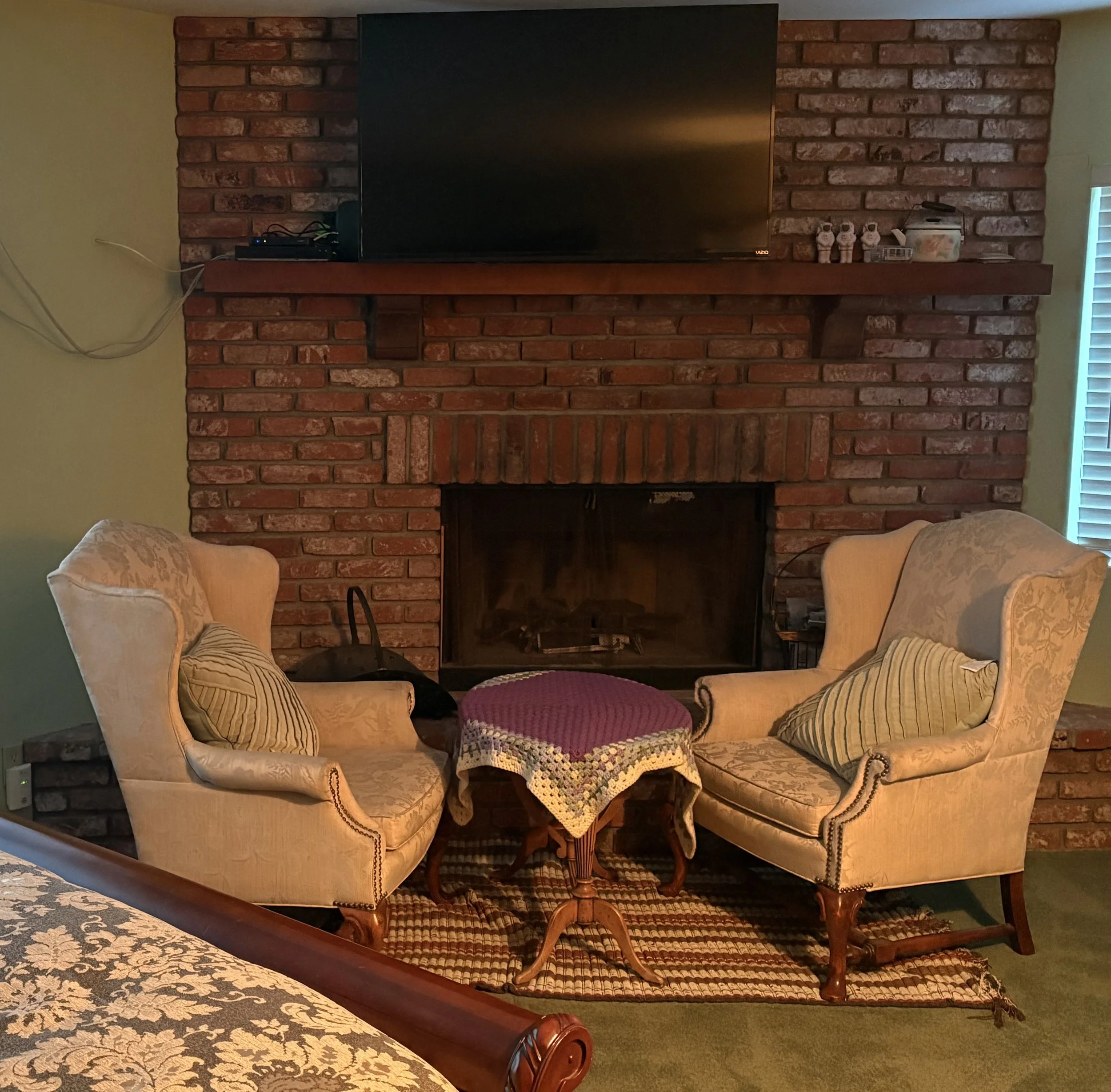 A cozy living room corner featuring a brick fireplace with a mounted flat-screen TV on the mantel. Two vintage-style armchairs with striped and patterned pillows flank a small wooden table covered with a crocheted doily. The room has warm, inviting decor and a green carpeted floor.