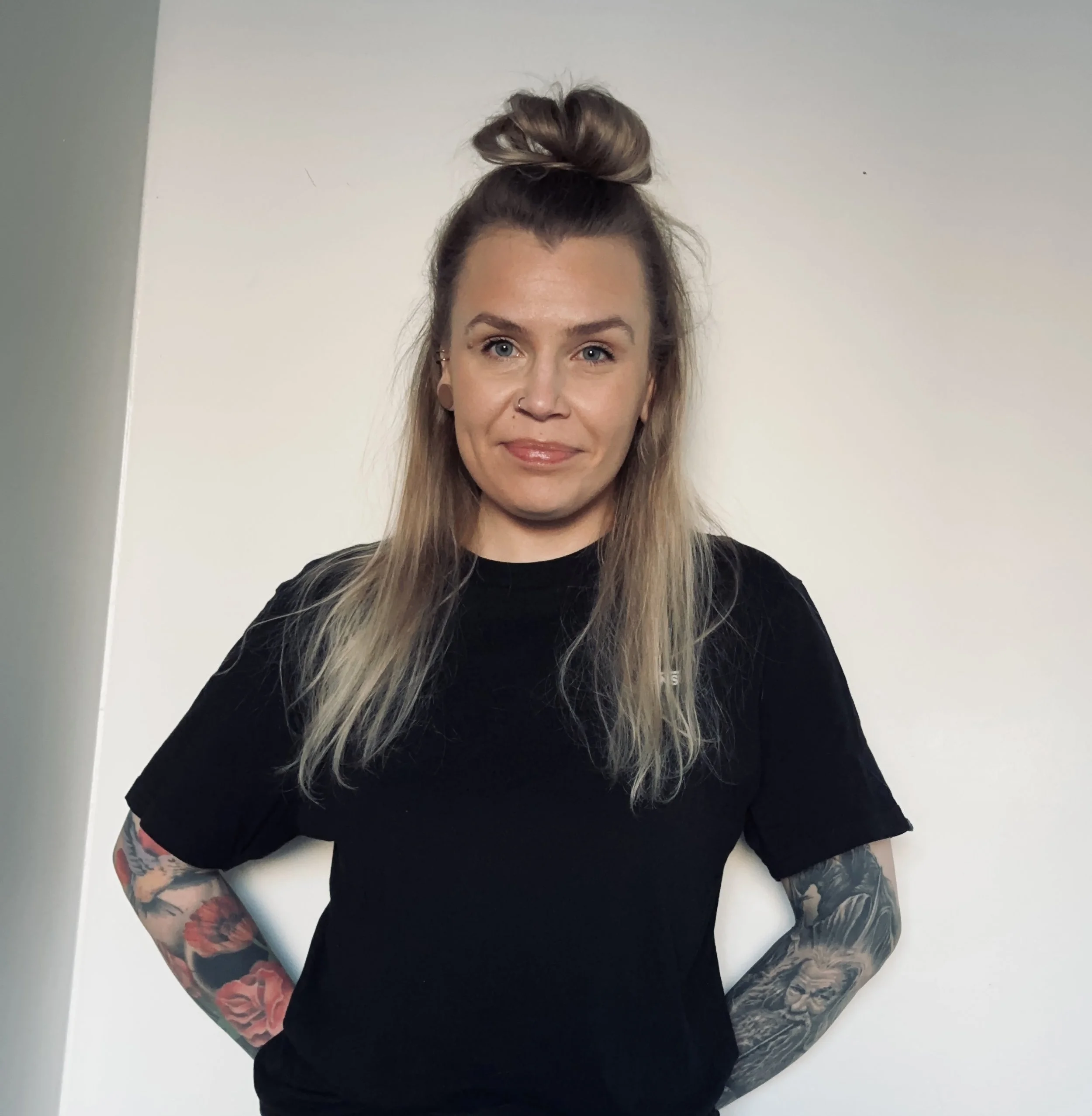 A woman with long blond hair styled in a top knot hairstyle, wearing a black t-shirt, standing against a plain white wall, with visible tattoos on both arms.