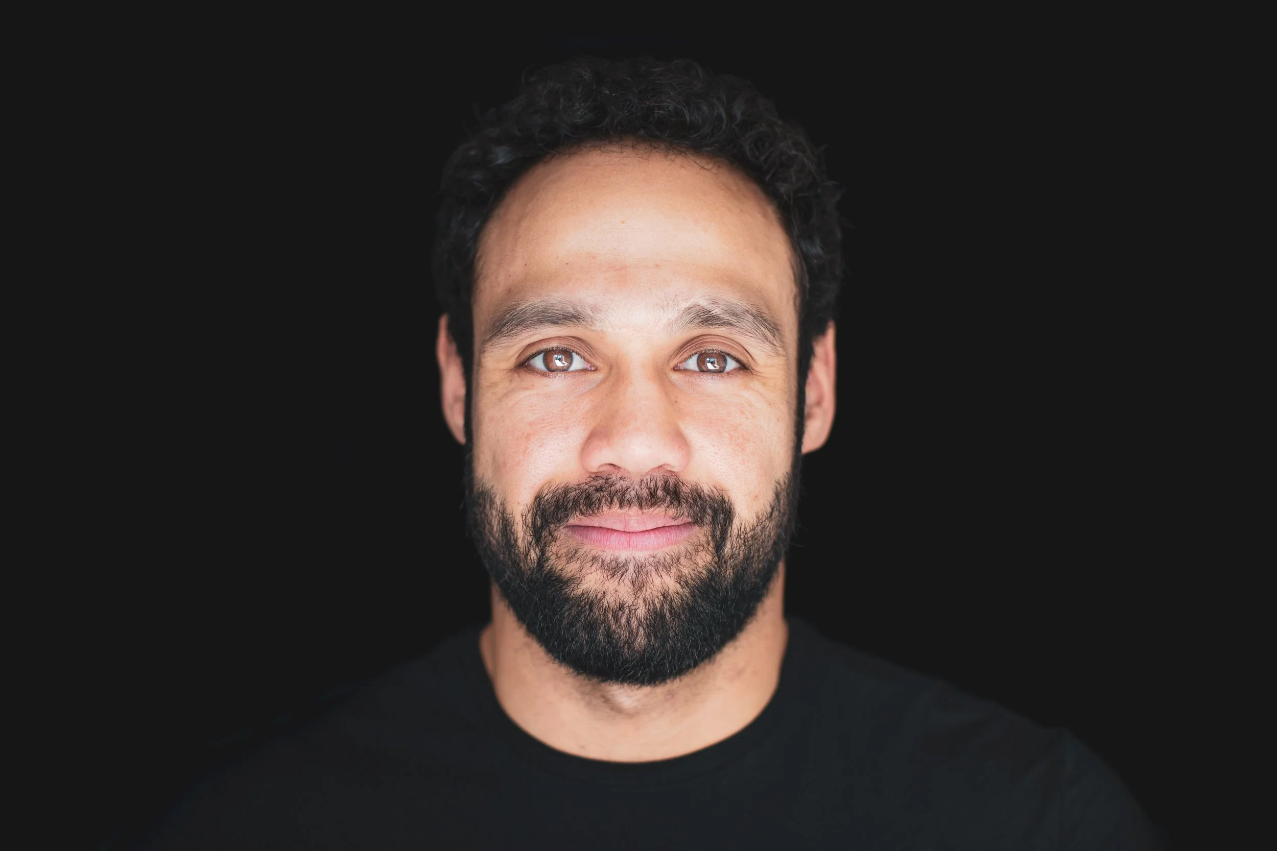 Portret van een man met een glimlach, donkere krullende haar en een volle baard, tegen een zwarte achtergrond.